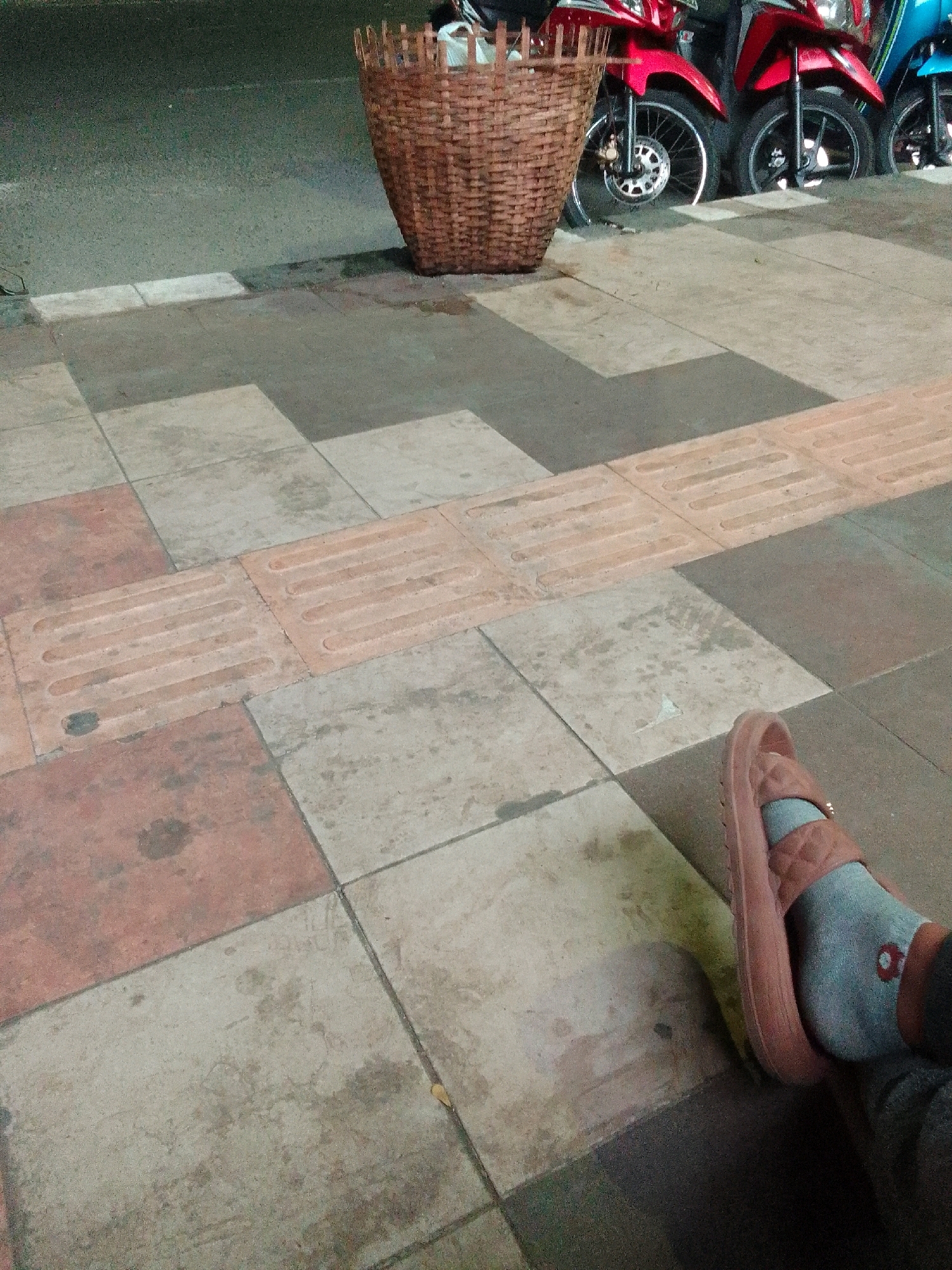The image depicts a street scene in Purbalingga, Indonesia, at ground level. In the foreground, a person's left foot is visible, wearing a light brown sandal and a gray sock with a small red floral design. The foot rests on a tiled sidewalk, characterized by a mix of reddish-brown and light gray square tiles. A section of tactile paving, designed to assist visually impaired pedestrians, is integrated into the sidewalk. Behind the sidewalk, a paved road surface is visible, and to the left, a portion of a woven basket is present. In the background, the wheels and lower sections of several motorcycles are parked, with the body of a red motorcycle being most prominent. The lighting suggests it is either early morning or evening.