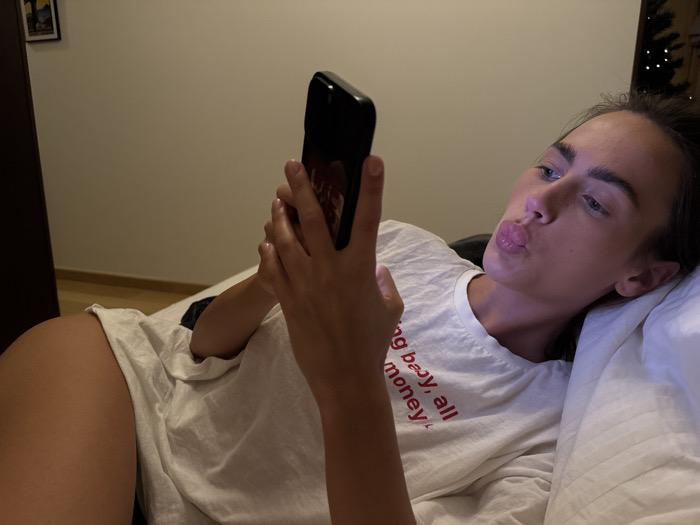 A young woman lies in bed, propped against pillows, holding a smartphone in her hand. She looks at the phone screen, pursing her lips in a "duck face" expression. She is wearing a white T-shirt with red lettering that reads "Young baby, all money." Part of her thigh is visible. The room is dimly lit, with a door and wall visible in the background. A Christmas tree is partially visible in the upper right corner.