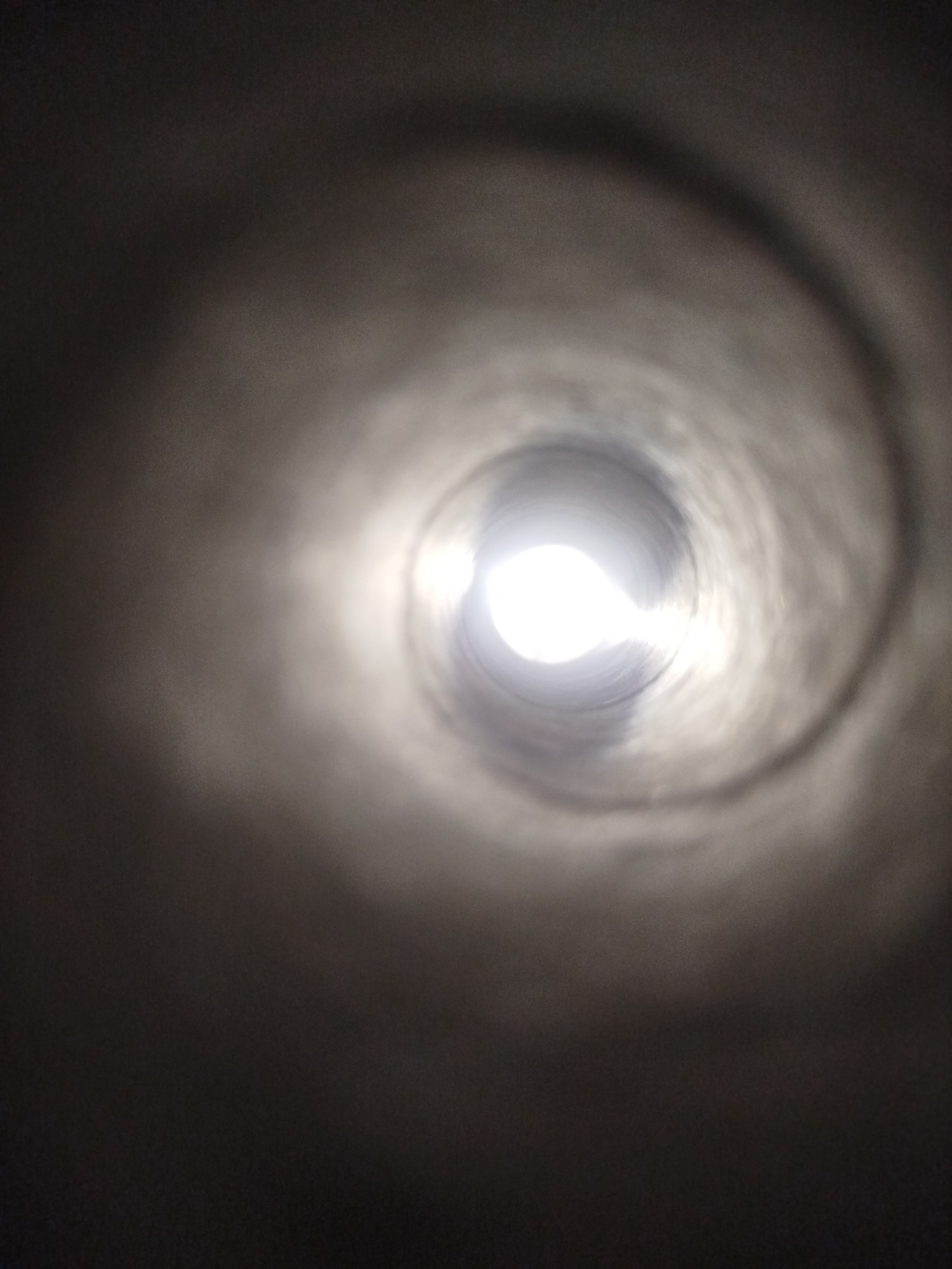 The image shows a view looking into a cylindrical tunnel or pipe. A bright light source is visible at the end of the tunnel, creating a strong contrast with the dark, textured walls of the pipe. The walls appear to be made of concrete or a similar material, with subtle variations in shade and texture that suggest a swirling pattern or a rough surface. The perspective gives the impression of looking into a deep, possibly endless, passage. There are no people, animals, or other identifiable objects within the frame. The scene does not provide any specific visual cues to pinpoint a location in Kremenchuk, Ukraine, or any other city.