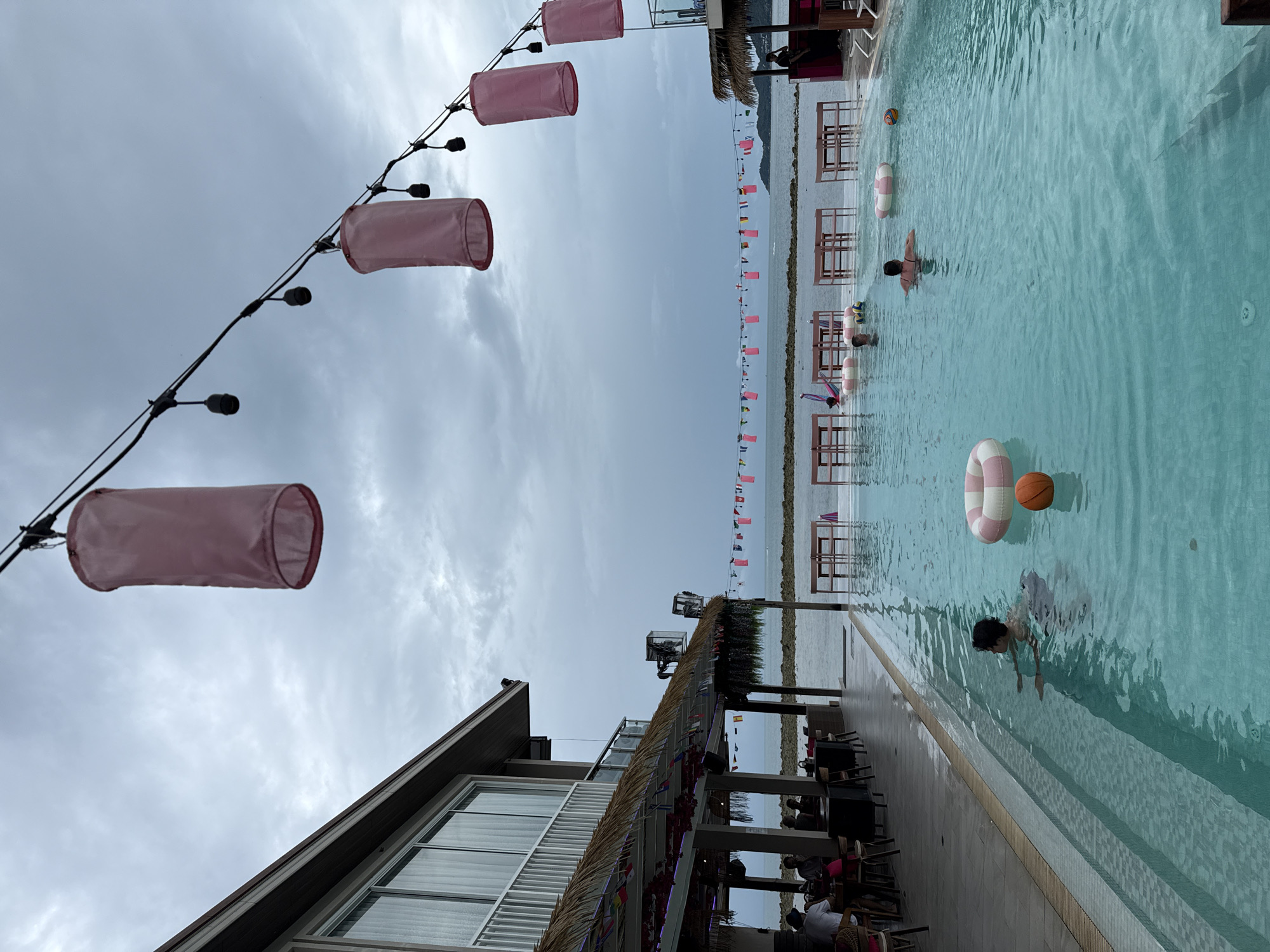 The image depicts an outdoor swimming pool area at what appears to be a beachfront establishment in the Ko Samui District of Thailand. The sky is overcast, with a muted, cloudy atmosphere suggesting it might be late afternoon or early evening.
In the foreground, a large, clear blue swimming pool dominates the scene. Several inflatable objects, including a pink and white striped ring and an orange basketball, are floating in the water. Two individuals are visible swimming in the pool: one person, likely male, is in the mid-ground, swimming towards the left side of the frame, and another person, possibly female, is in the right mid-ground, facing away from the camera.
Adjacent to the pool, there is a paved walkway and then a building with a thatched roof over an outdoor dining or seating area. This area has tables and chairs set up. Flags of various colors are strung along the edge of the thatched roof and also in a long line above the water, stretching towards the horizon. In the background, the sea is visible, with a distant shoreline or island on the right.
Above the pool and building, a string of black electrical wire holds several large, pink fabric lanterns, which are a prominent decorative element. The building itself has a modern architectural style with large windows on an upper level. The overall scene suggests a relaxed, tropical resort or restaurant setting.