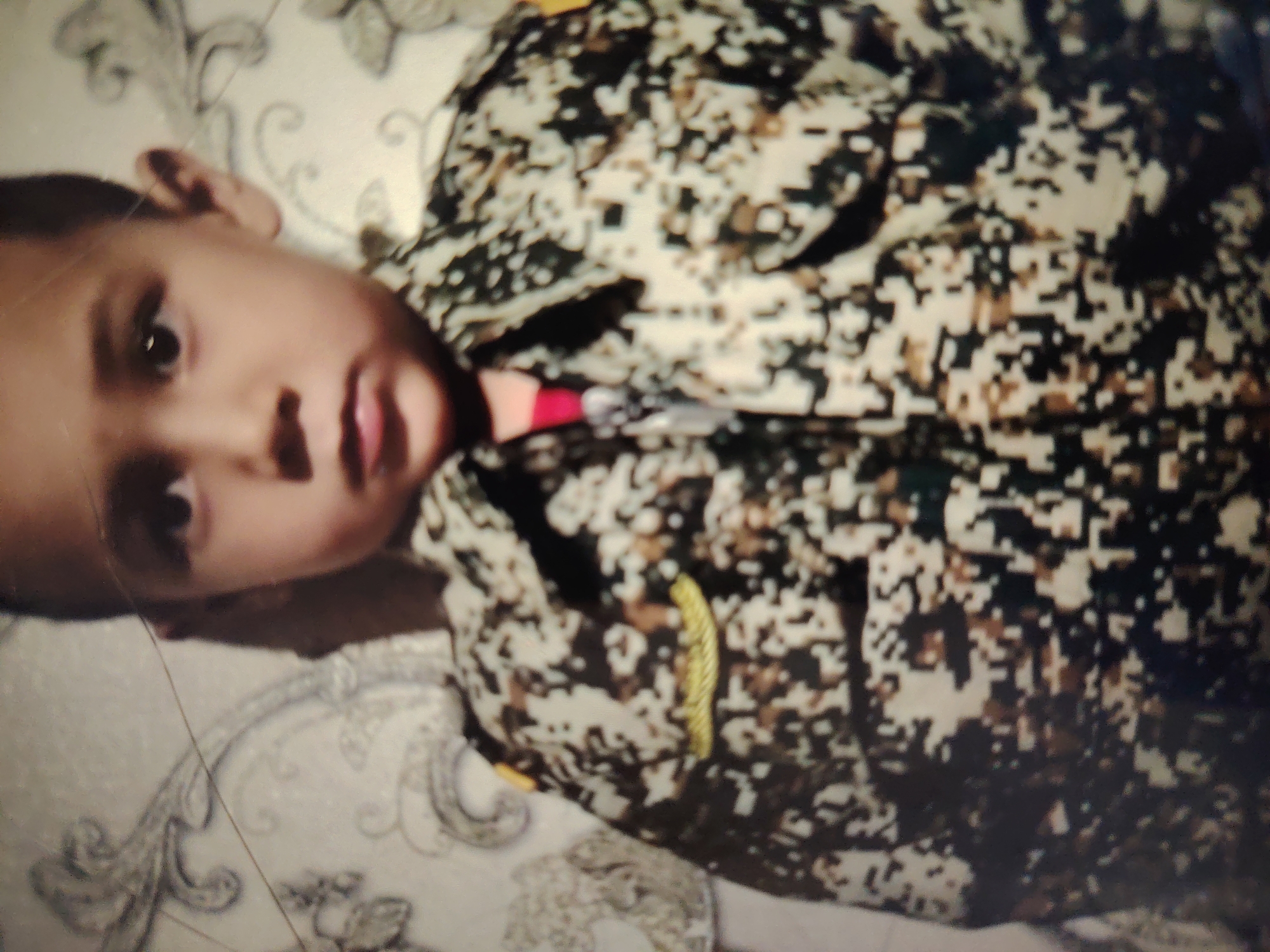 The image shows a close-up, angled view of a young child lying down. The child is wearing a camouflage-patterned shirt with pixelated brown, black, and white colors. A portion of a red object is visible near the collar of the shirt. The child's head is tilted, with their face partially visible, showing an eye and mouth. The background appears to be a light-colored fabric with a faint, ornate pattern. There are no discernible features that would indicate the location in Bahshitpa, Uzbekistan. The image quality is low and blurry.