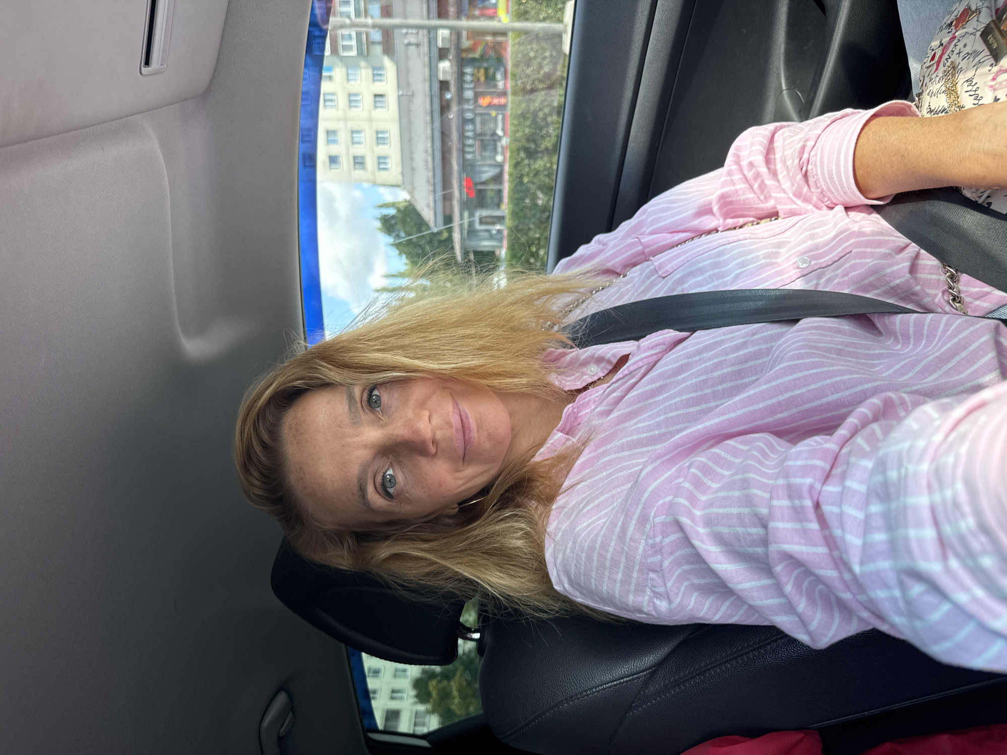 A woman with blonde hair and blue eyes, wearing a pink and white striped shirt and a seatbelt, is seated in the driver's seat of a car. The car's interior is visible, with a black leather seat and the car's ceiling. Outside the car, a building with multiple windows and a sign that reads "KEBAB OT MEGO" is visible, indicating the location is likely a city street in Sinyavino, Russia. The time of day appears to be daytime, with sunlight streaming in.