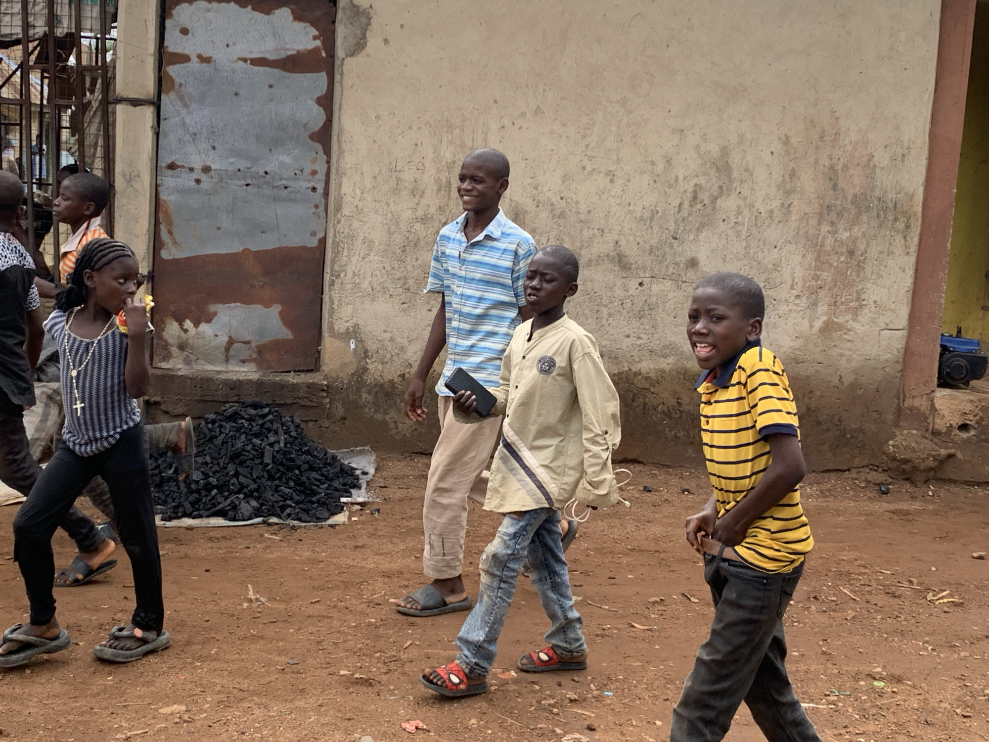 Akwanga, Nigeria: A group of children walk past a building with a heavily rusted metal door. A girl walks to the left of a pile of charcoal and is holding food in her hand. A boy in a blue striped shirt holds a dark-colored cell phone in his left hand. Another boy walks to his right, carrying a white charging cable. To the right, a boy in a yellow-striped shirt walks toward the right side of the frame. The ground is a red-brown dirt.