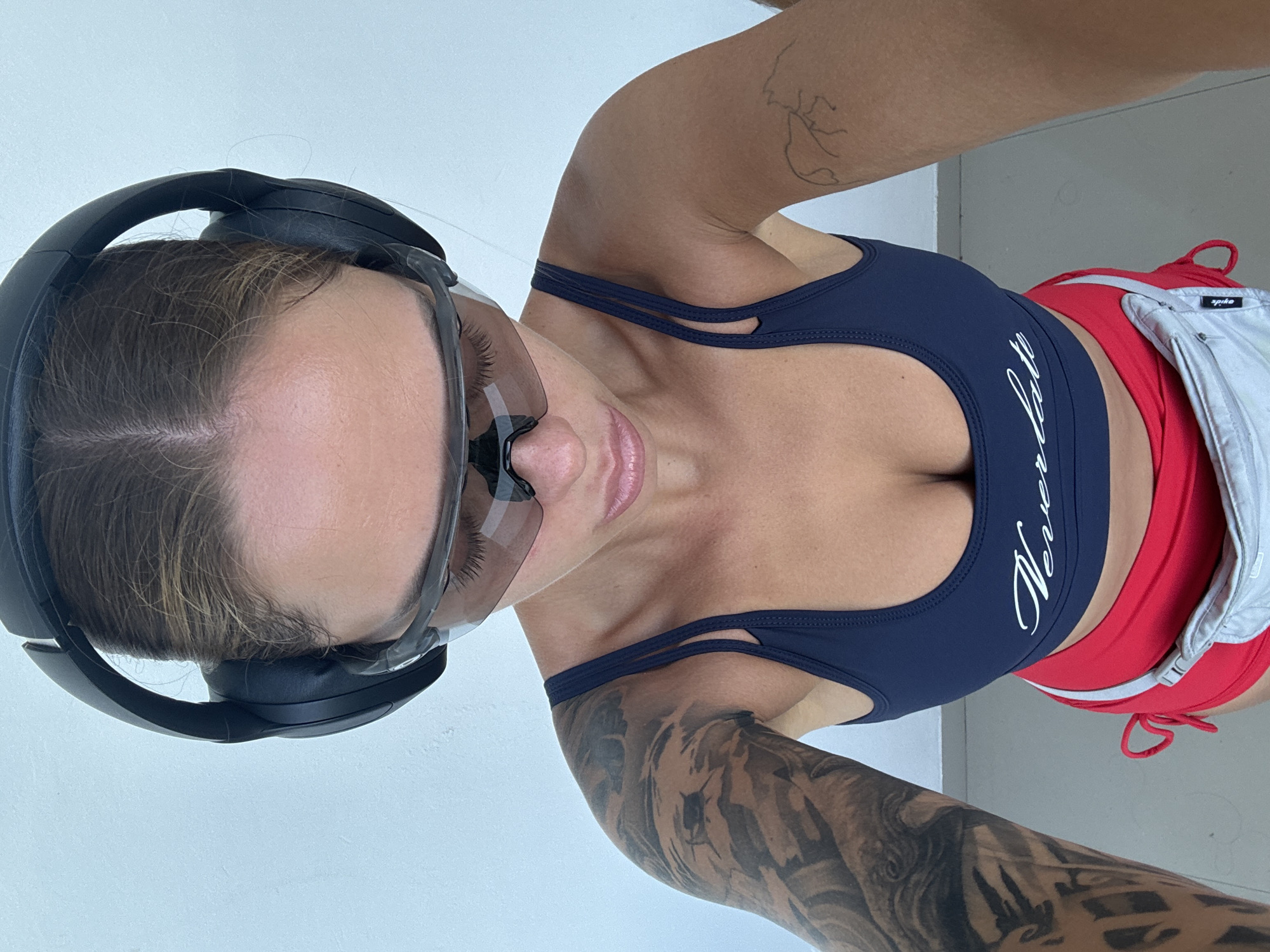 A selfie from a first-person perspective, showing a woman in activewear. She wears a navy-blue sports bra with white lettering, red shorts, and a gray waist pack. The woman has dark hair and is wearing sunglasses and headphones. The background appears to be a white wall and a gray floor.
This image does not contain sufficient information to determine a specific location within North Kuta, Indonesia, or confirm it is located there. The indoor setting lacks distinctive geographical or cultural cues that would indicate the location.