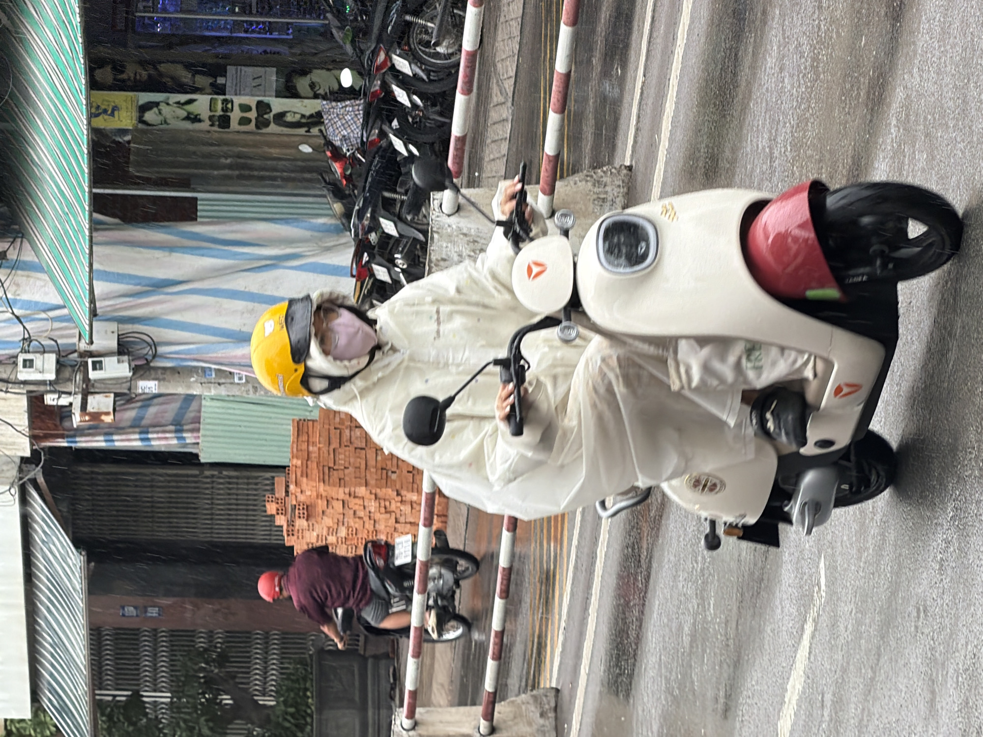 In Nha Trang, Vietnam, on a rainy day, a person wearing a white raincoat and a yellow helmet is riding a white and red scooter. The rider has on a pink mask. Another person on a motorcycle with a red helmet is visible in the background, moving away from the camera. The street is wet and reflects the surroundings. Buildings with awnings and some construction materials, specifically stacks of orange bricks, are visible along the side of the street. There are also several parked motorcycles in front of a building. The overall scene depicts urban traffic in inclement weather.