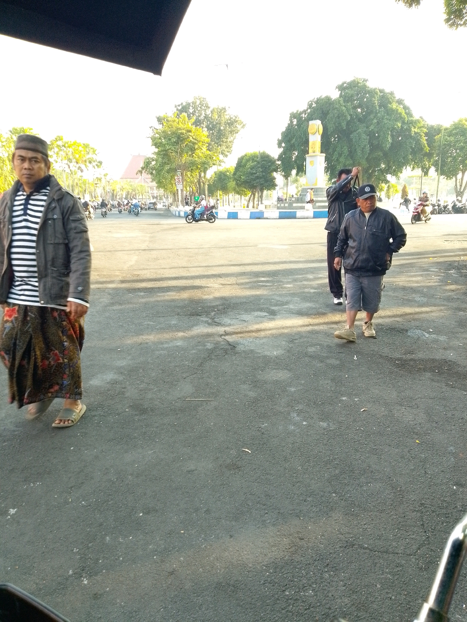 The image depicts an outdoor scene with several individuals and vehicles on a paved surface, likely a street or public square. In the foreground, on the left, a person wearing a dark jacket over a striped shirt and a patterned sarong walks towards the camera. On the right, another person wearing a dark jacket, a light-colored shirt, gray shorts, and a baseball cap walks away from the camera. In the middle ground, various motorcycles are visible, some with riders, suggesting traffic or a gathering. A tall, light-colored monument with yellow accents stands in the background, surrounded by trees. The environment appears to be a public area with a clear sky and sunlight casting shadows. Based on the presence of the monument and the overall urban setting, this location is likely the Tugu Pancasila (Pancasila Monument) in Jember, Indonesia.