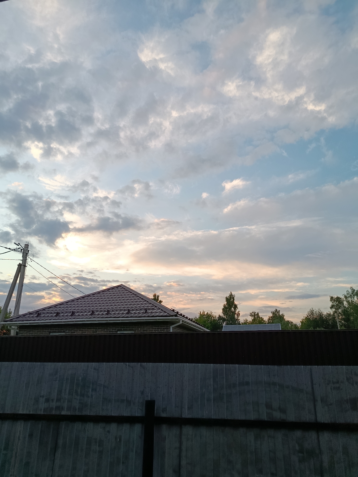 The image depicts a cloudy sky with a hint of sunset colors over a residential area in Vereya, Russia. In the foreground, a dark metal fence runs horizontally across the frame. Behind the fence, a dark brown corrugated metal fence is visible, followed by the dark tiled roof of a house. To the left, a utility pole with wires is present. The background features a line of trees and more houses, with the sky dominating the upper portion of the image. The lighting suggests it is either sunrise or sunset.
