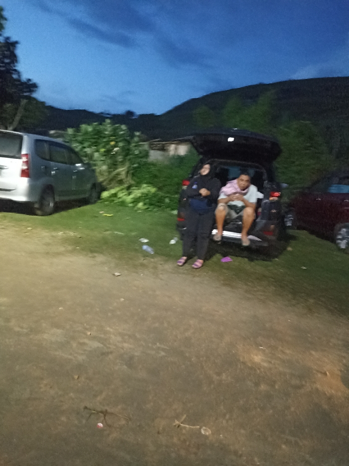 This image captures an outdoor scene at dusk or early night in Kecamatan Pujut, Indonesia. The sky above is a deep twilight blue, with the silhouette of distant hills or mountains visible against it.
In the center-right of the frame, two individuals are present near a dark-colored SUV or minivan. The vehicle's trunk is open, serving as a resting spot. A woman, wearing a dark hijab, a dark long-sleeved top, and dark pants, stands beside the open trunk, smiling towards the camera. Inside the trunk, a man is seated, wearing a light-colored t-shirt and shorts, looking relaxed.
The ground beneath them is an unpaved, earthy surface, illuminated by an unseen artificial light source, creating a warm glow. Several empty plastic bottles are scattered on the ground near the dark car, suggesting recent consumption. To the left, a silver minivan or SUV is parked, partially visible. On the far right, a portion of a red car can also be seen. The background features lush green vegetation and bushes, with a small, indistinct structure faintly visible behind them. The scene suggests a casual stop or rest during travel. No visible text is present in the image.