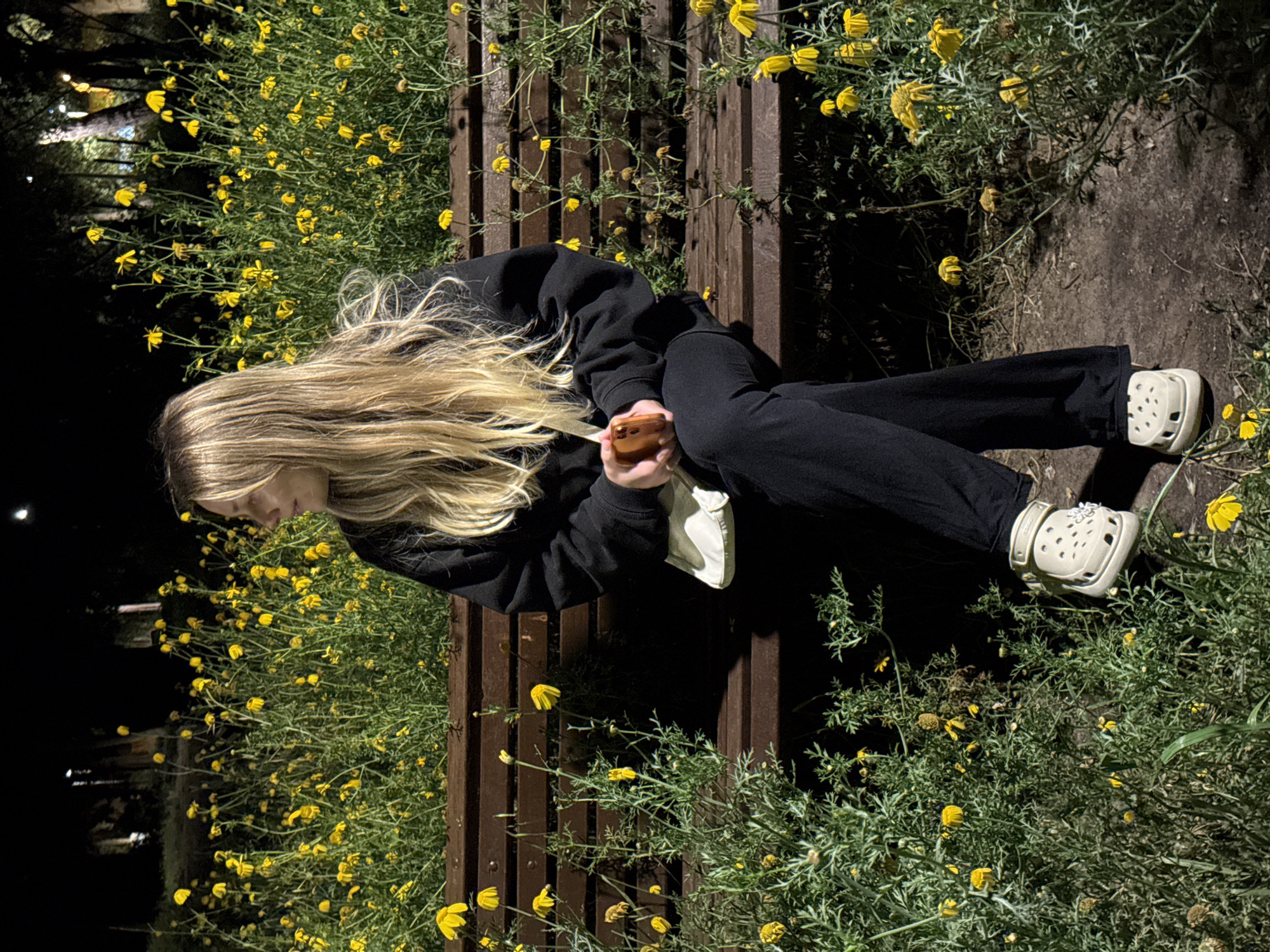 A young woman with long, blonde hair sits on a dark brown wooden bench in an outdoor setting at night. She wears a black hoodie and black pants, with light-colored Crocs on her feet. She is holding a smartphone in her hands. Yellow flowers surround the bench, growing both behind and in front of it, suggesting a garden or park-like environment. The setting is dark, with a light source visible in the background on the left side. The scene is likely in Tel Aviv, Israel.