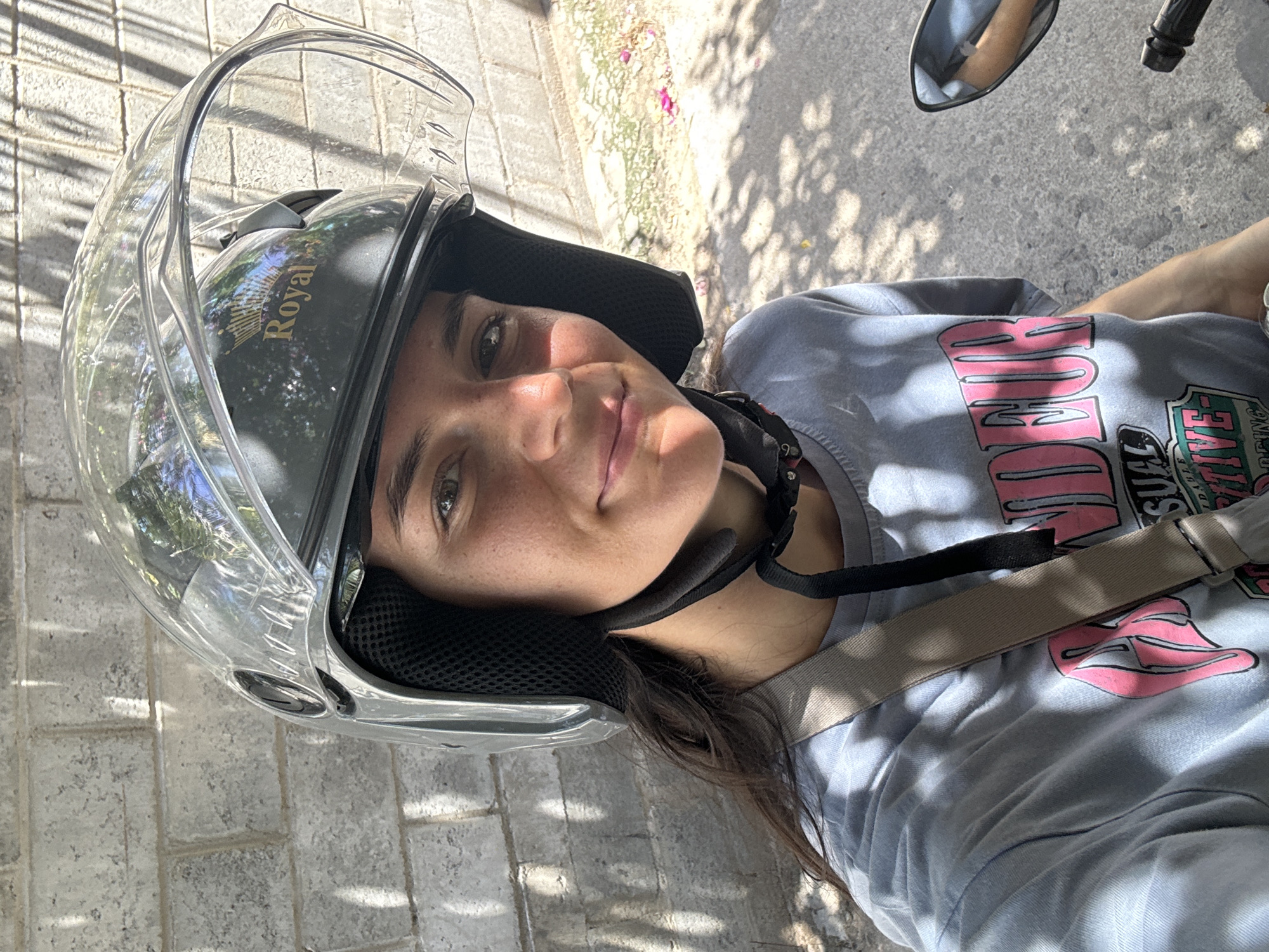 A young woman wearing a gray helmet with a clear visor, a light blue t-shirt with pink accents, and a beige strap across her chest is seen in a selfie. She appears to be on a motorcycle, as a side mirror is partially visible at the top right. The photo is taken outdoors in Mũi Né, Vietnam, indicated by the Royal helmet and typical architecture of the region, with a brick pavement beneath and shadows from trees and foliage in the background.