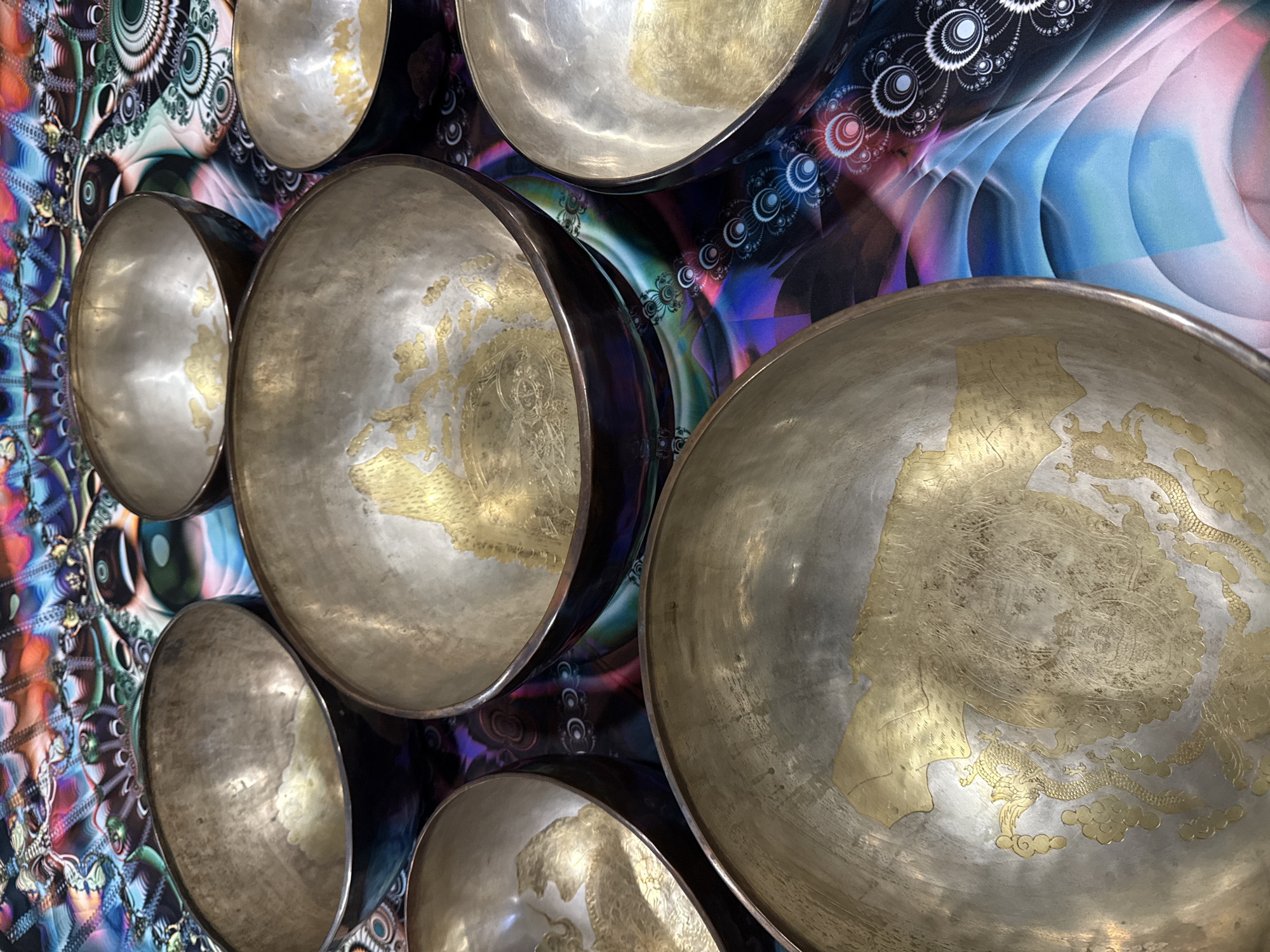 The image shows a collection of singing bowls mounted on a wall in Tashkent, Uzbekistan. The bowls are silver with ornate gold designs. The wall behind them is decorated with a colorful pattern of geometric shapes and swirls. There are no people visible.