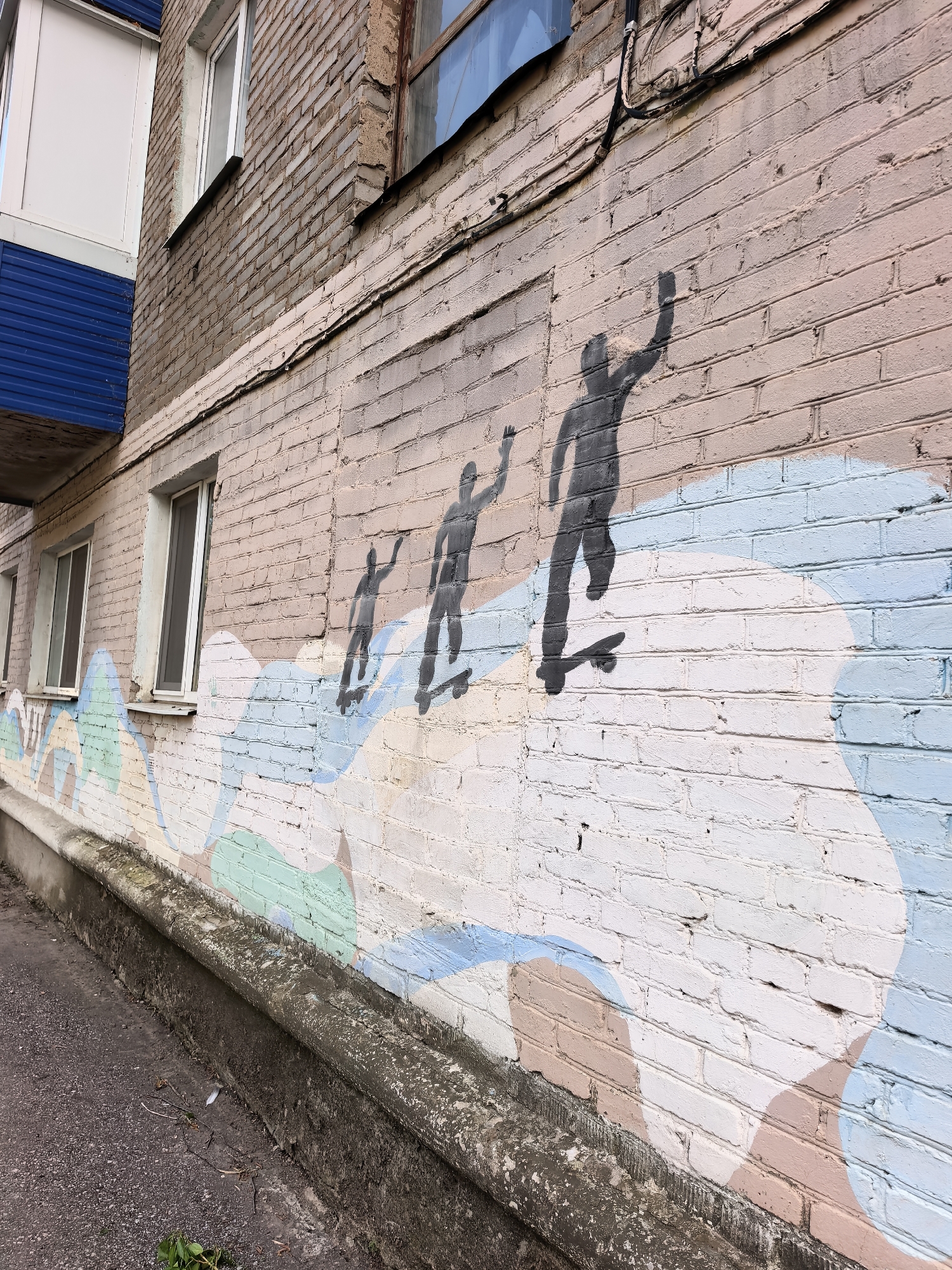 A street-level view shows a brick building's exterior wall in Sterlitamak, Russia, featuring a mural of three dark silhouette figures riding skateboards on a wave-like painted design. The figures are spaced progressively further apart, with their arms raised, suggesting movement. The wall is painted in shades of pink, white, light blue, and green, creating a stylized wave beneath the skateboarders. Windows are visible on the building. The ground is asphalt and a concrete ledge runs along the base of the wall. A few pieces of greenery are visible near the edge of the asphalt. Cables are visible on the brick wall.