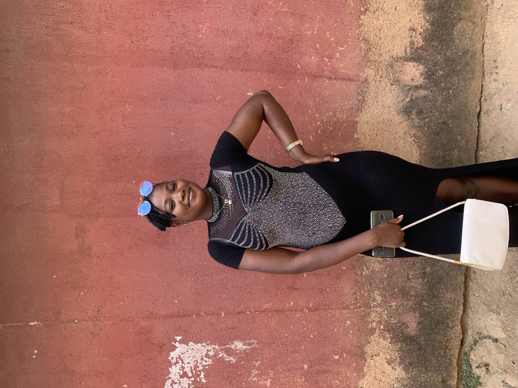 A young woman stands against a weathered wall painted in shades of red and tan in Jos, Nigeria. She is wearing a black dress with a glittering silver pattern on the bodice and a slit on the side. Her hair is styled in braids, and she wears round, blue-tinted sunglasses perched on her head. She smiles, her head slightly tilted. A gold bracelet adorns her wrist. Her right hand is on her hip, and her left hand holds a smartphone and the strap of a small, white purse. The wall is marked with uneven patches and textures, suggesting age and exposure to the elements. The ground at the bottom of the frame is concrete with cracks and patches of greenery. The overall impression is a casual portrait in an urban setting.