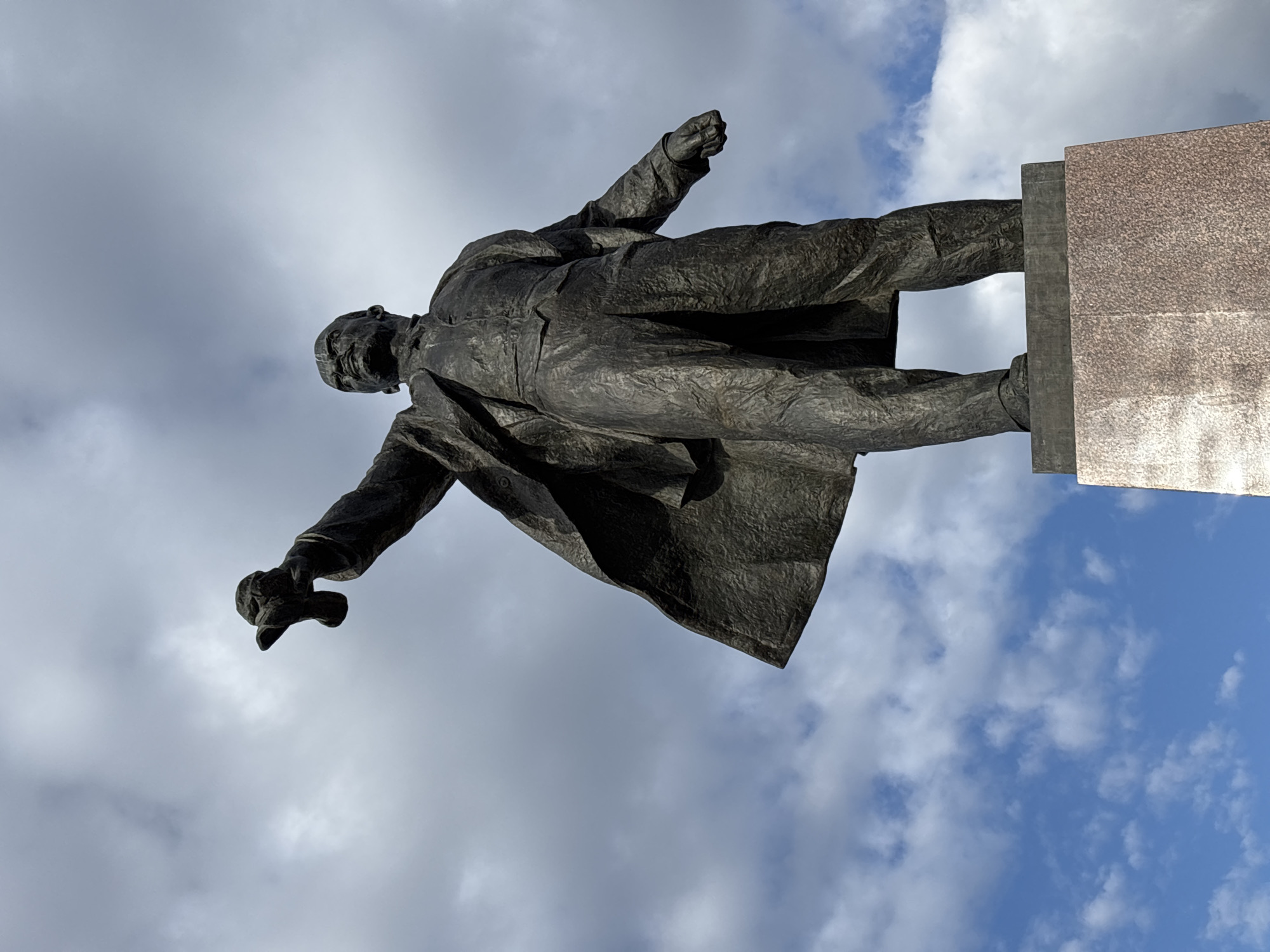 A low-angle shot of a bronze statue set against a cloudy blue sky. The statue depicts a man, likely Vladimir Lenin, in a suit with a long coat, standing on a base of red granite. He has his right fist clenched in the air, and his left hand holds a rolled-up paper. The statue is likely the Lenin monument at Moskovskaya Square in Saint Petersburg, Russia.