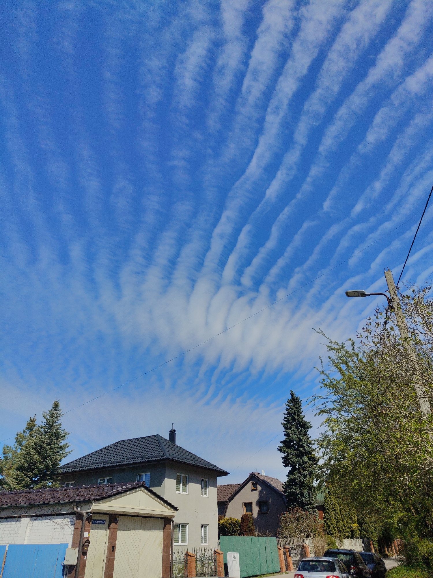 The image shows a residential street in Unknown, Unknown. Several houses are visible, some with trees in front of them. There are cars parked on the street. A street lamp is on the right side of the image. The sky is blue with long, parallel bands of clouds.