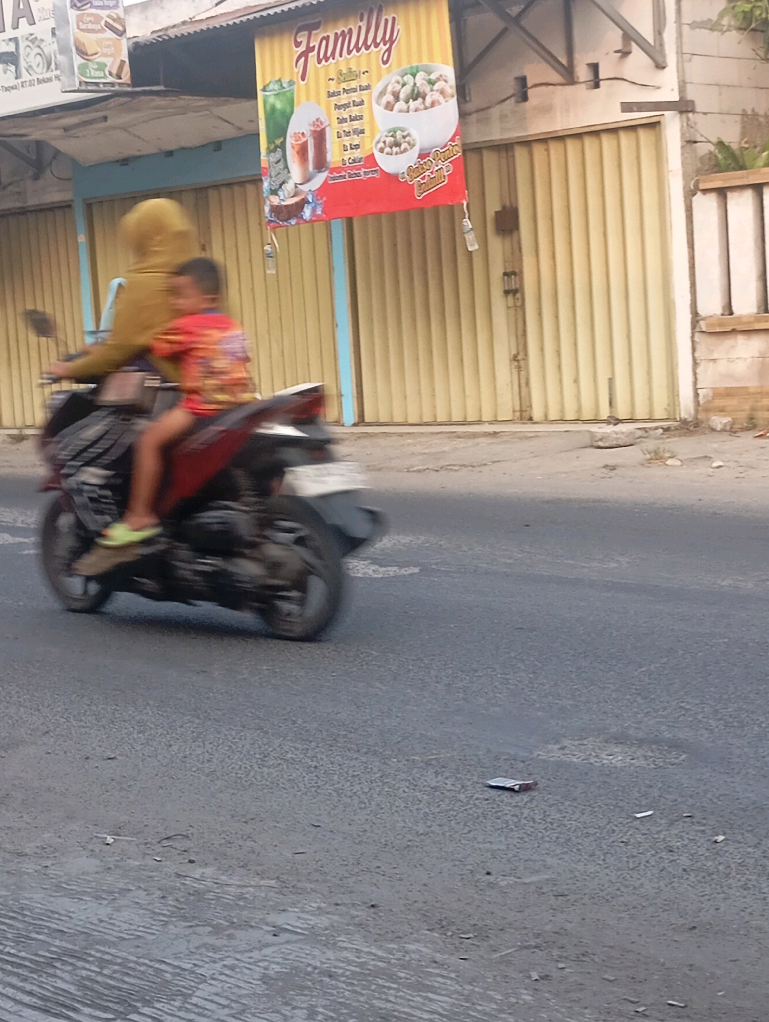 An adult wearing a yellow hijab and a child in a red shirt are riding a motorcycle on a paved road in Babelan, Indonesia. The adult is operating the motorcycle, while the child is seated behind them. The motorcycle is in motion, as indicated by the blur in the image. In the background, a building with a yellow roll-up door and a banner advertising "Family" with images of food and drinks is visible. The environment appears to be an urban or semi-urban setting, with the road in the foreground and the building and banner behind.