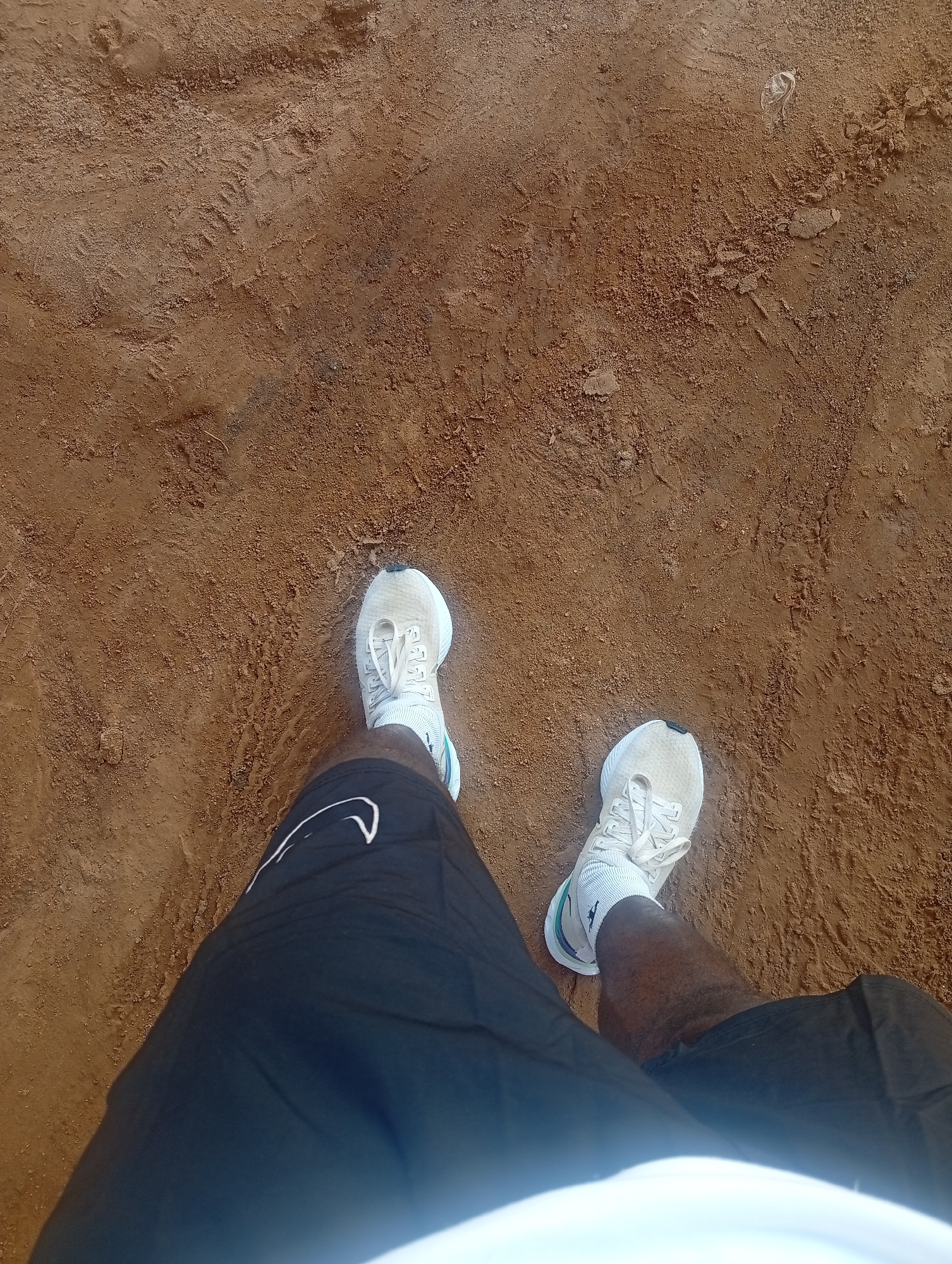 The image shows a person's feet wearing white athletic shoes and black shorts standing on a dirt ground. The ground is brown and appears to be dry, with some footprints visible. The lighting suggests it is daytime. The location is the city of Jos, Nigeria.