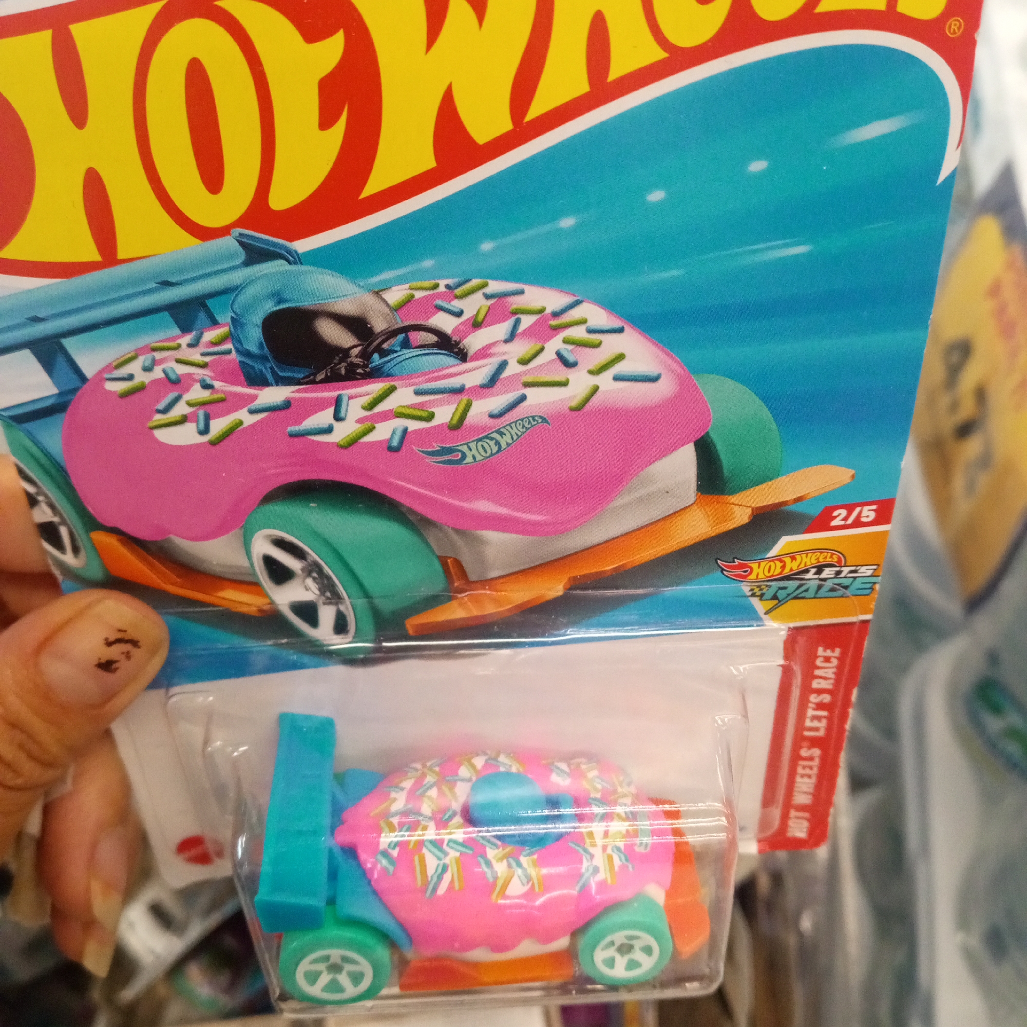 The image shows a Hot Wheels toy car packaging. The packaging is primarily red and yellow with a blue background. The main focus is a Hot Wheels car designed to look like a pink donut with sprinkles. It has turquoise wheels and orange accents. A portion of a person's hand with dark marks on a fingernail is visible in the lower left corner, holding the packaging. The background shows a blurred store environment. The context suggests this is a retail product display. The location of El Limon, Venezuela is not present in the visual information of the image.