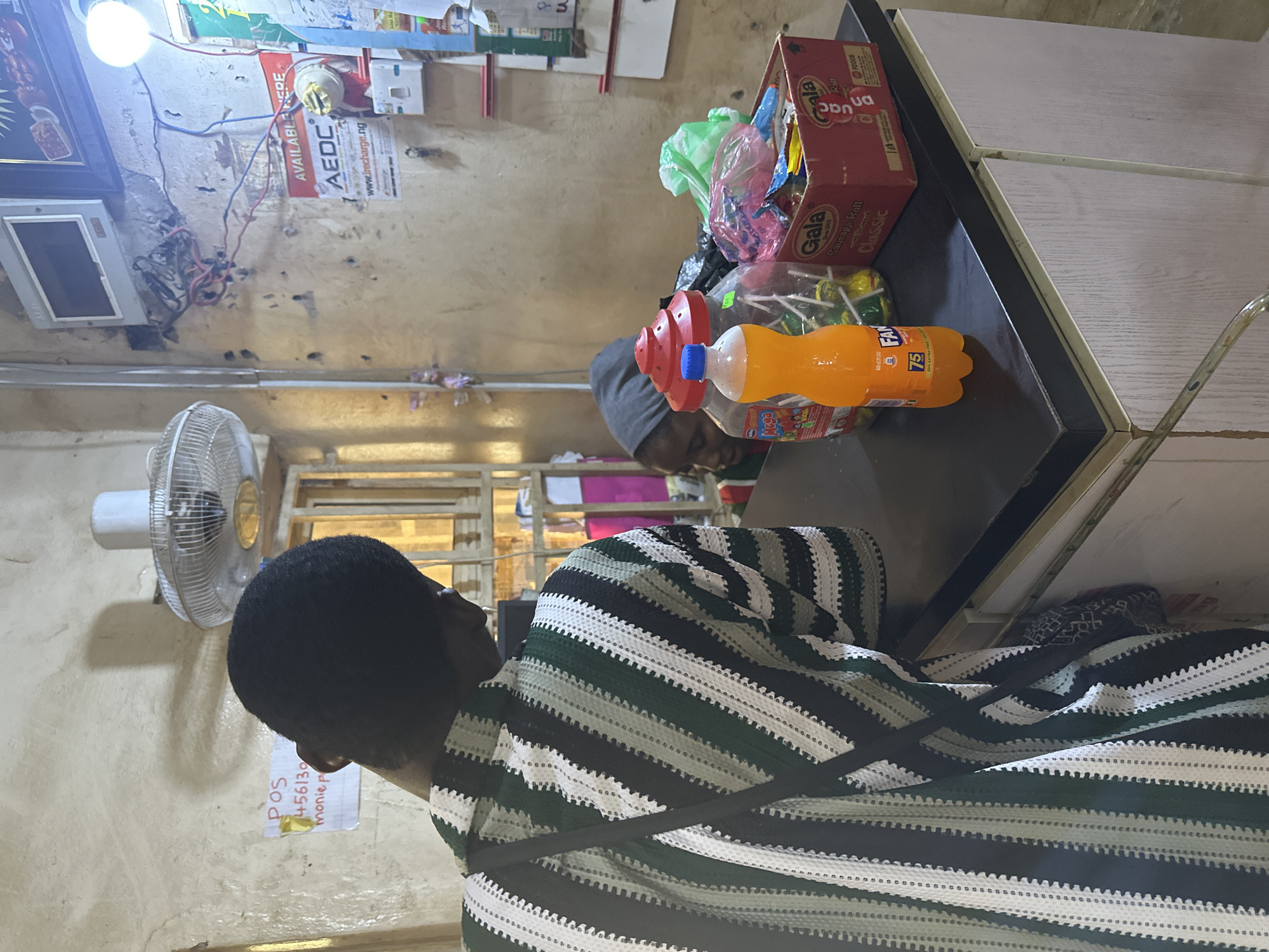 The image depicts a young person wearing a green, white, and black striped garment, with their head bowed and facing away from the camera. They are positioned in what appears to be a small shop or convenience store, suggested by the presence of merchandise like a box of "Gala Sausage Roll" and a bottle of orange soda labeled "Mega Fanta".
In the background, a fan is visible on the left, and a bright lightbulb illuminates the ceiling area, which is adorned with various posters and electrical wiring. A notice on the wall reads "POS 456130 monie p" and another poster mentions "AEDC" and "www.irecharge.ng". The setting seems to be indoors, with a worn and cluttered aesthetic, possibly indicating a busy or well-used establishment. The overall impression is that of a candid snapshot of daily life within a Nigerian retail environment. The lighting suggests it might be daytime, though the artificial light source is prominent. No specific event is depicted; the scene appears to capture a moment of pause or engagement with an unseen task.
