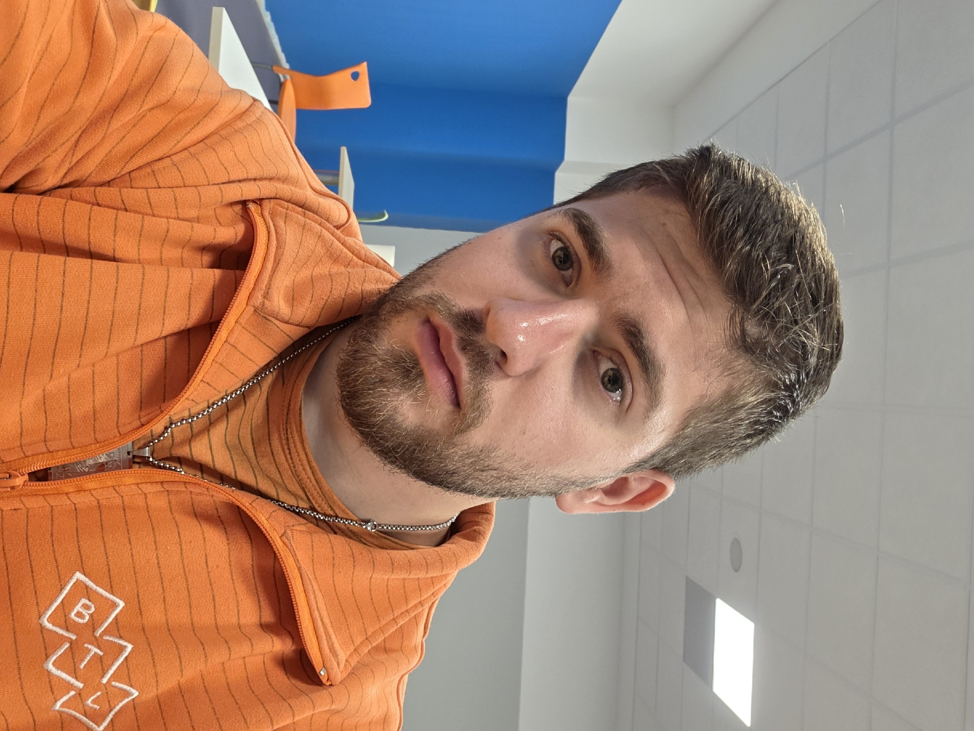 A close-up photograph shows a man with short brown hair, light brown eyes, and a beard. He is wearing an orange, vertically striped hooded jacket with a white logo on the left side of the chest. A silver-colored chain necklace is visible around his neck. The background is composed of a blue ceiling element, white walls with rectangular lighting panels, and other blurred objects. The location is unknown, and there is no information to suggest it is in Plovdiv, Bulgaria.