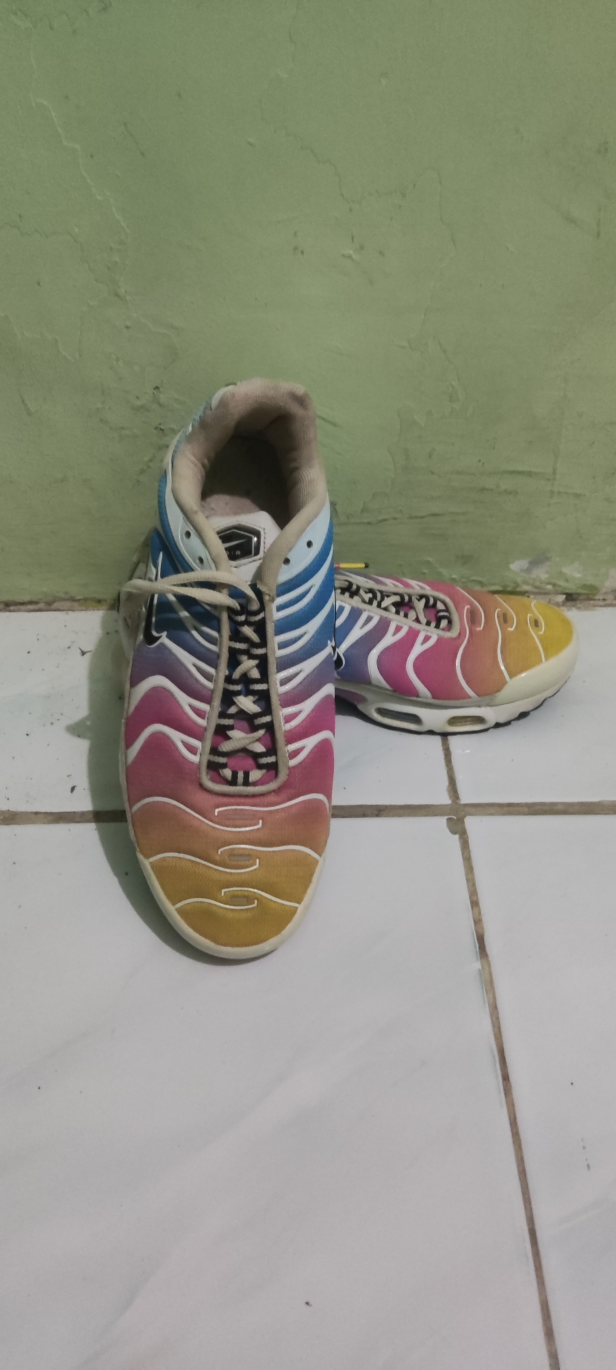 A pair of colorful Nike Air Max Plus sneakers are displayed on a tiled floor in front of a pale green wall. The left shoe is positioned upright, facing the viewer, with its laces tied. The right shoe is placed next to it, angled slightly away. The sneakers feature a gradient color scheme, transitioning from blue at the top to pink and yellow towards the toe. The background wall appears slightly textured and shows some signs of wear. The floor is covered in white square tiles with visible grout lines. The scene is static and appears to be indoors. The location is not explicitly stated to be in Gebang, Indonesia.