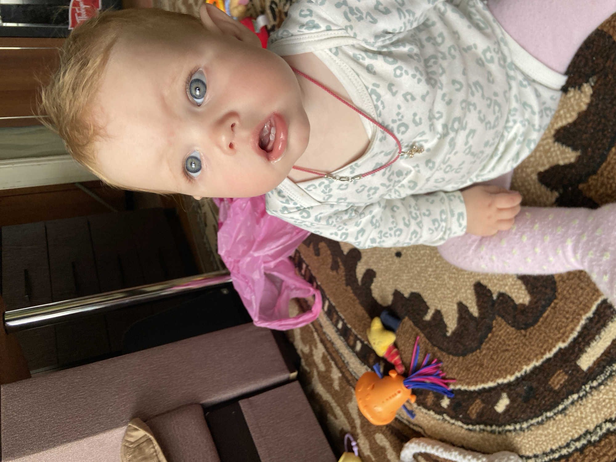 This media file captures an infant lying on a patterned rug in an indoor setting in Фоки, Russia. The baby, with light reddish-blonde hair and wide, striking blue eyes, looks directly towards the camera with a curious expression, mouth slightly open, revealing two small lower teeth.
The infant is dressed in a white long-sleeved onesie adorned with a light grey animal print pattern, paired with pink polka-dotted leggings. A thin pink cord necklace with a small metallic charm hangs around the baby's neck.
Around the baby, the floor is covered by a textured rug featuring an abstract pattern in shades of brown, beige, and black. Scattered on the rug are several colorful toys, including an orange and blue one with frills, and a small yellow one. A crinkled pink plastic bag lies near the baby's side. In the background, sections of dark wooden furniture or cabinetry are visible, along with a metallic bar. A blurry red object or label, possibly with faint text such as "Virginia," is partially visible in the upper left background. The overall scene suggests a casual moment in a home environment.