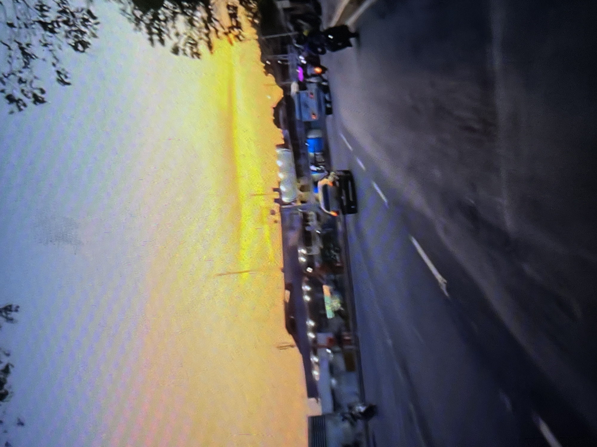 A street scene in Tinaquillo, Venezuela, at dusk. The sky transitions from blue to yellow. Cars and motorcycles are parked along the side of the road and driving down it. Buildings line the road. There is a tree in the foreground.