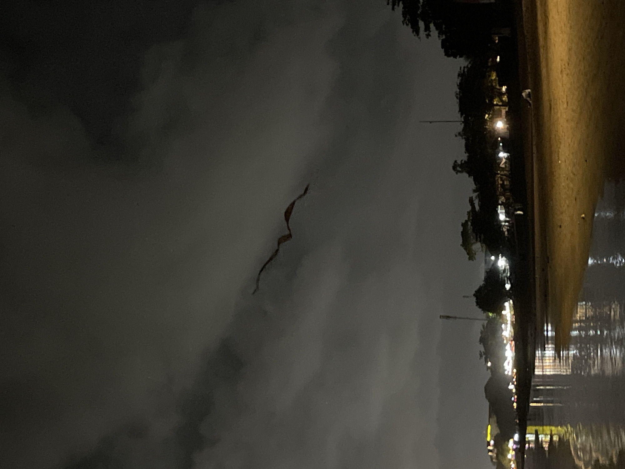 A nighttime aerial view of a beach in Kuta Selatan, Indonesia shows a dark, cloudy sky. A kite shaped like a dragon is flying in the sky. In the background, a beach is visible with palm trees and lights from buildings. The water reflects the lights of the city, and there is a sandy beach with some people walking.