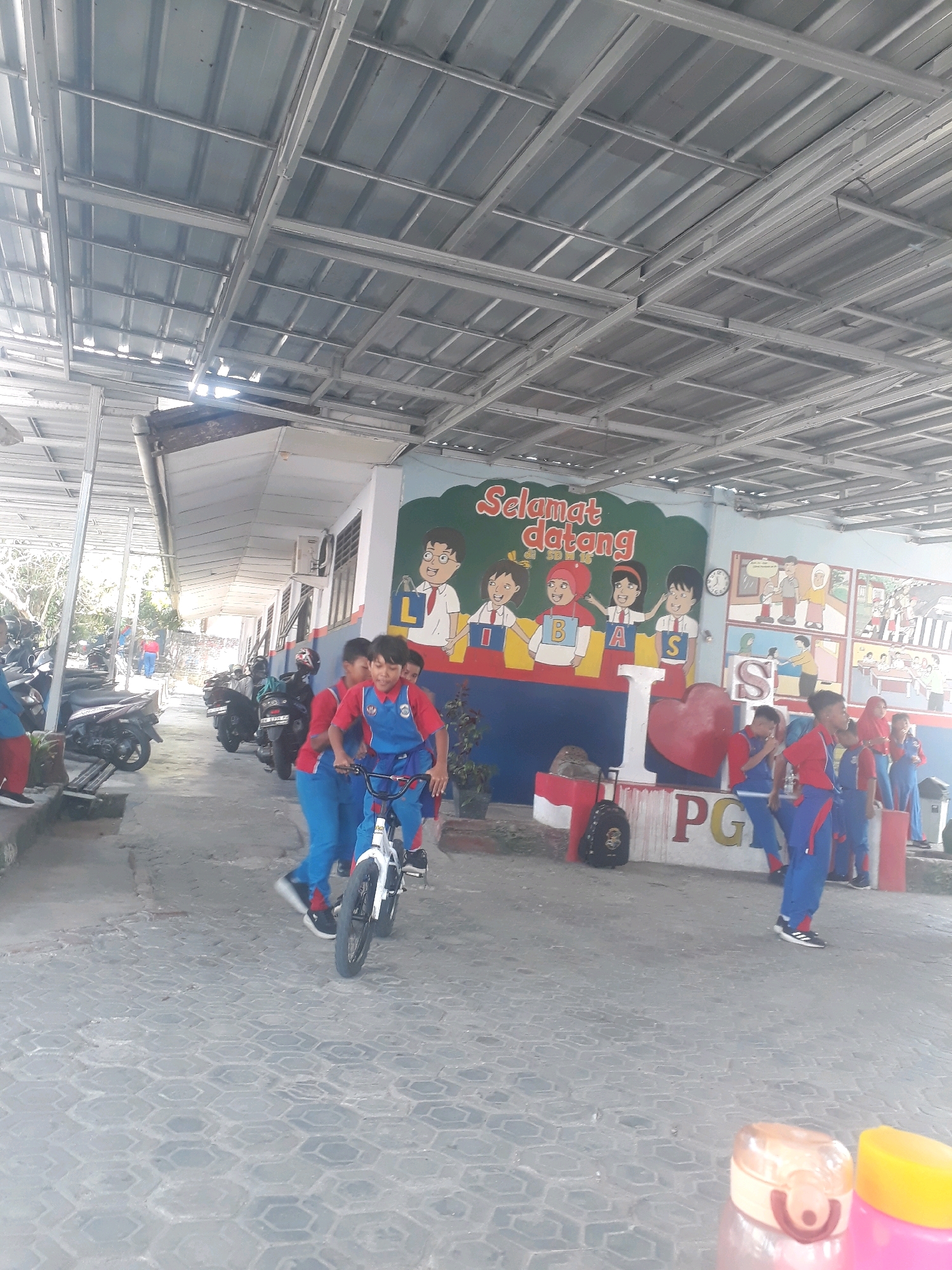 The image shows a group of schoolchildren in uniform, some riding a bicycle, in an outdoor area that appears to be a school or community center in Pangkalpinang, Indonesia. The children are wearing blue and red uniforms, with some also wearing red headscarves. The background features a building with a mural on its wall that says "Selamat Datang" (Welcome) and depicts children. There are motorcycles parked along the side of the paved walkway. The area is covered by a metal roof structure. In the foreground, a backpack and two water bottles are visible on the pavement. The overall scene suggests a lively and casual school environment.