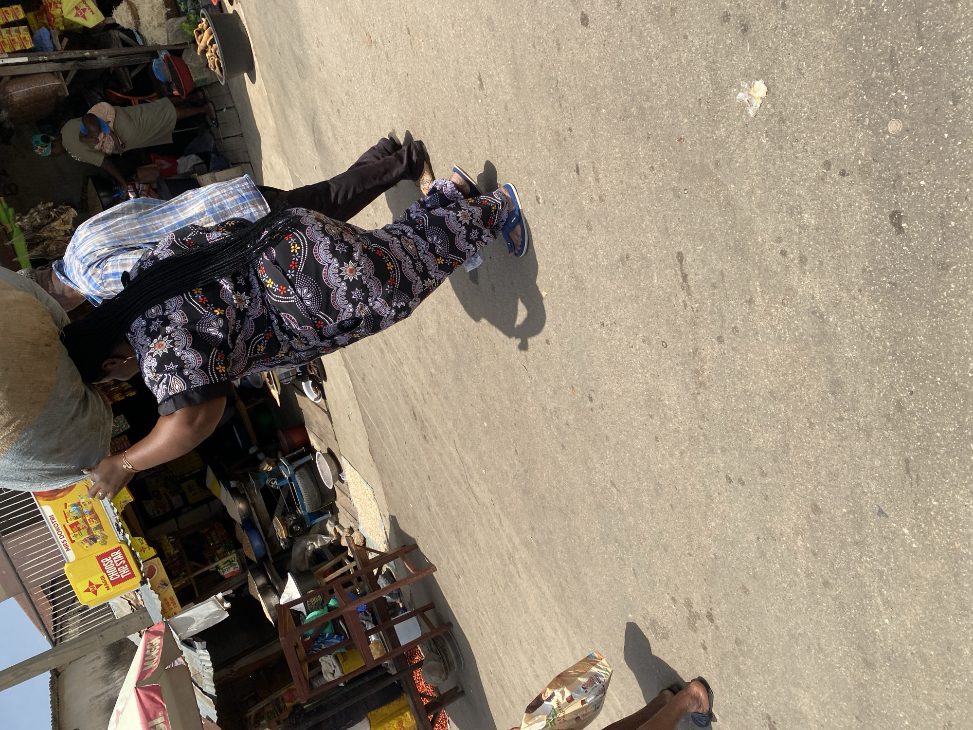 The image depicts a bustling street scene in Jos, Nigeria, under a clear, sunny sky. A woman, positioned in the foreground and walking away from the camera, carries a large, light-colored sack on her head. She is wearing a vibrantly patterned, dark-colored dress and blue flip-flops. Beside her, a man walks to her left, wearing a blue and white checkered shirt and dark trousers. In the background, a market stall displays various goods, including produce and packaged items. A woman in the background is seen carrying a child on her back. The ground is paved with asphalt, and there are some scattered pieces of debris. The overall impression is one of everyday activity in a Nigerian urban setting.
