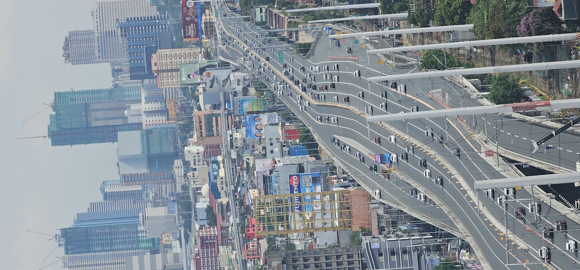 An aerial, full shot captures a sprawling urban landscape in Makati, Philippines, dominated by a multi-lane elevated highway filled with vehicles. The highway curves and splits, with traffic flowing in both directions. On either side of the highway, the cityscape is dense with buildings of varying heights, from low-rise structures to towering skyscrapers, some of which are still under construction, indicated by scaffolding and cranes. Billboards are visible along the sides of the highway and on top of some buildings. The sky is overcast, suggesting a hazy or possibly rainy day. The foreground shows the concrete supports of the elevated highway, with some construction work visible to the right, including dirt piles and barriers. The overall scene depicts a busy, modern metropolitan area undergoing continuous development.