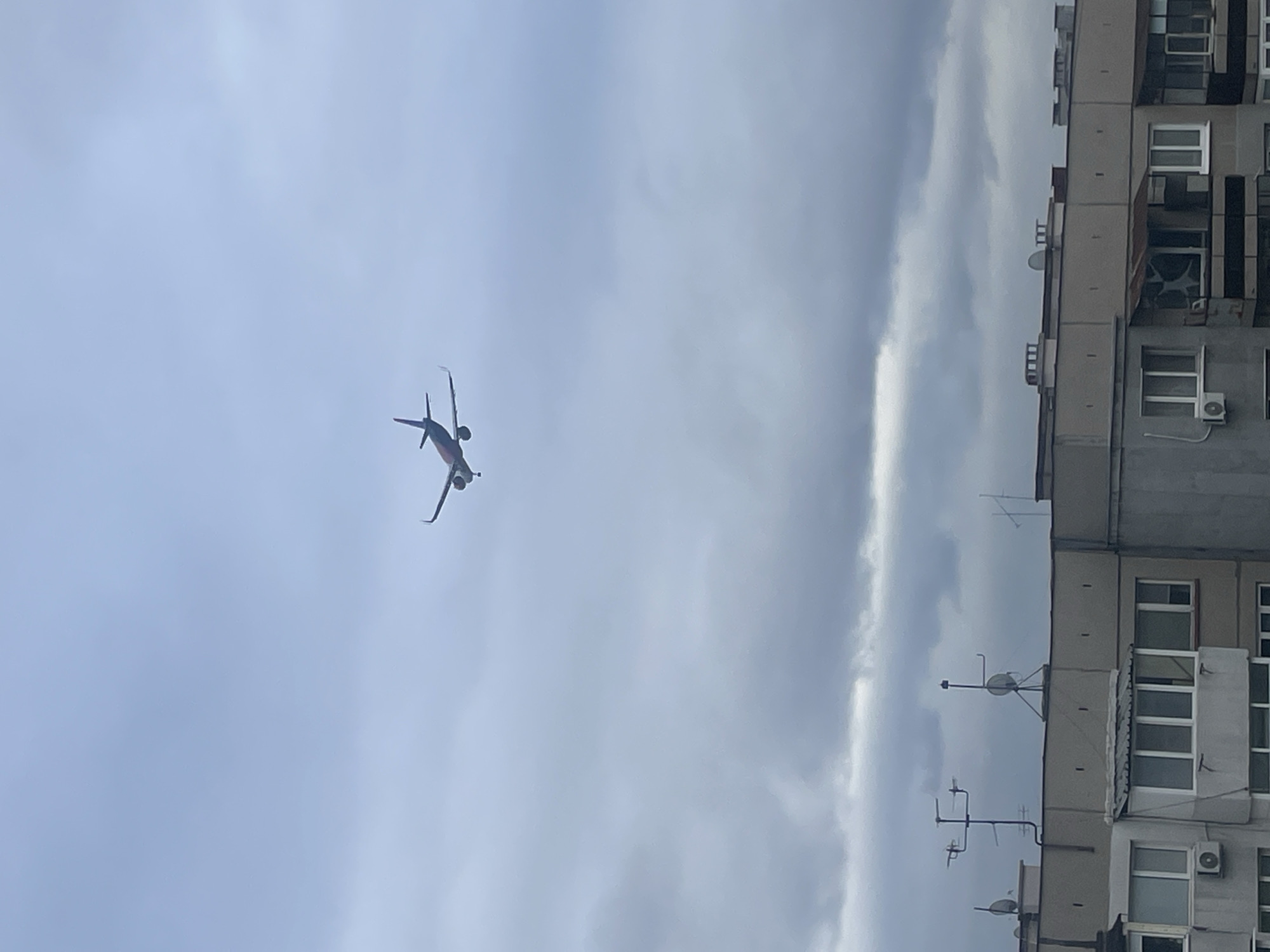 The image shows an airplane flying in the sky above a building. The sky is cloudy. The building is a multi-story residential building. The location is most likely the city of Sofia, Bulgaria.