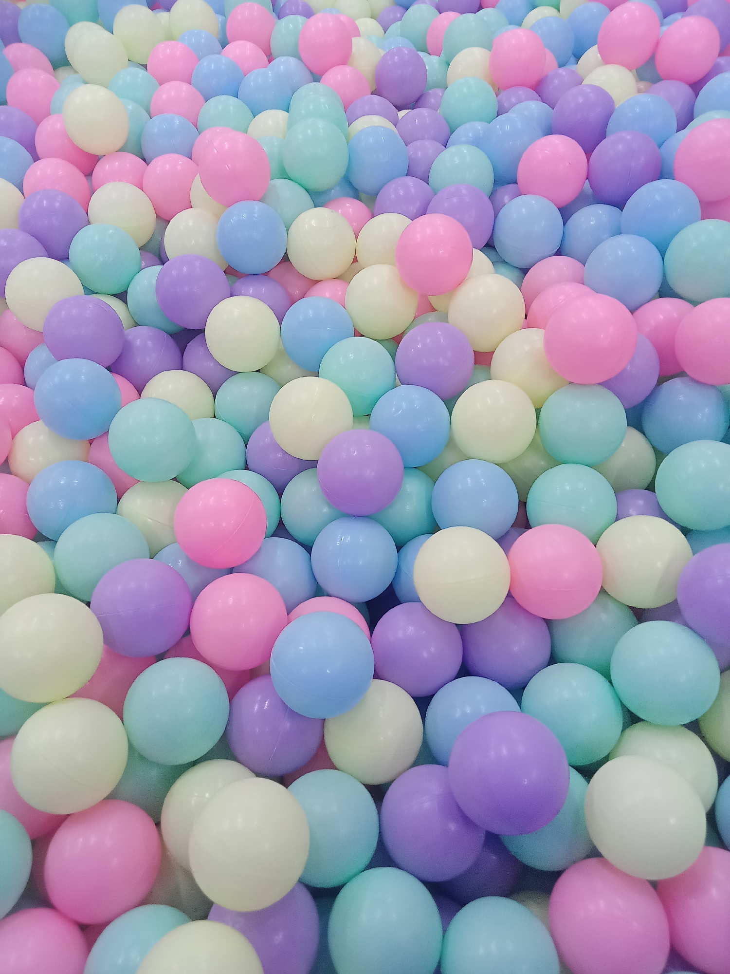 The image is a close-up, top-down view of a large collection of pastel-colored plastic balls in various shades of pink, purple, blue, and yellow. The balls are of similar size and are densely packed, creating a soft, textured surface with no discernible pattern. The lighting is bright and even, suggesting an indoor setting, possibly a ball pit in a children's play area. There are no people or animals visible in the image, nor any specific landmarks that would indicate a precise location within Boyolali, Indonesia. The scene is entirely focused on the abstract arrangement of the colored balls.
