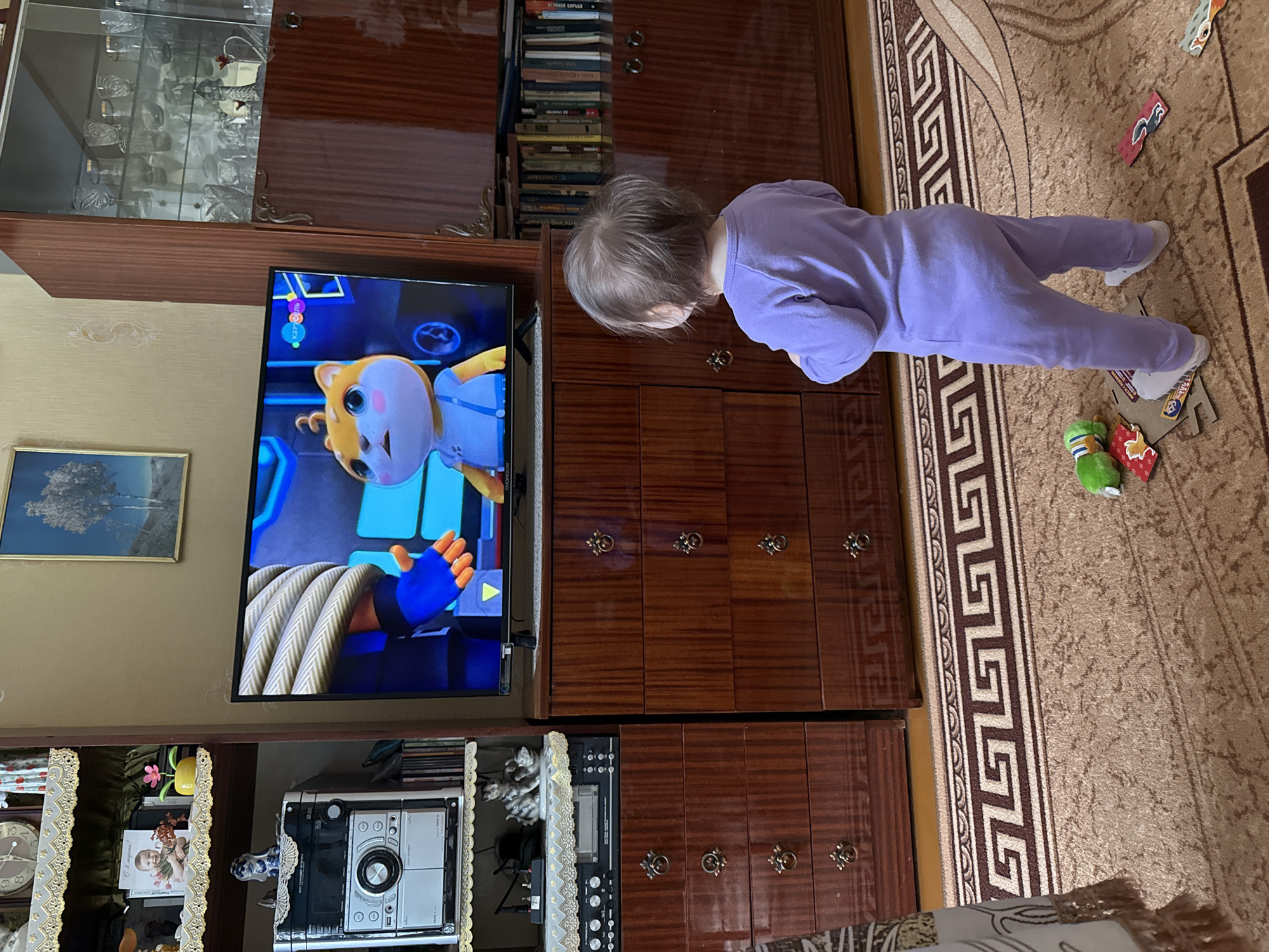 A child in a lavender jumpsuit is watching a cartoon on a television set. The TV is set inside a brown wooden cabinet. To the left of the TV, is a clear glass cabinet filled with glassware. There is a picture hanging on the wall behind the TV. Below the TV is a stereo system. The child is standing on a brown carpet with a geometric pattern and puzzle pieces scattered on the floor. The location is likely Vyshegor, Russia, based on the provided information.