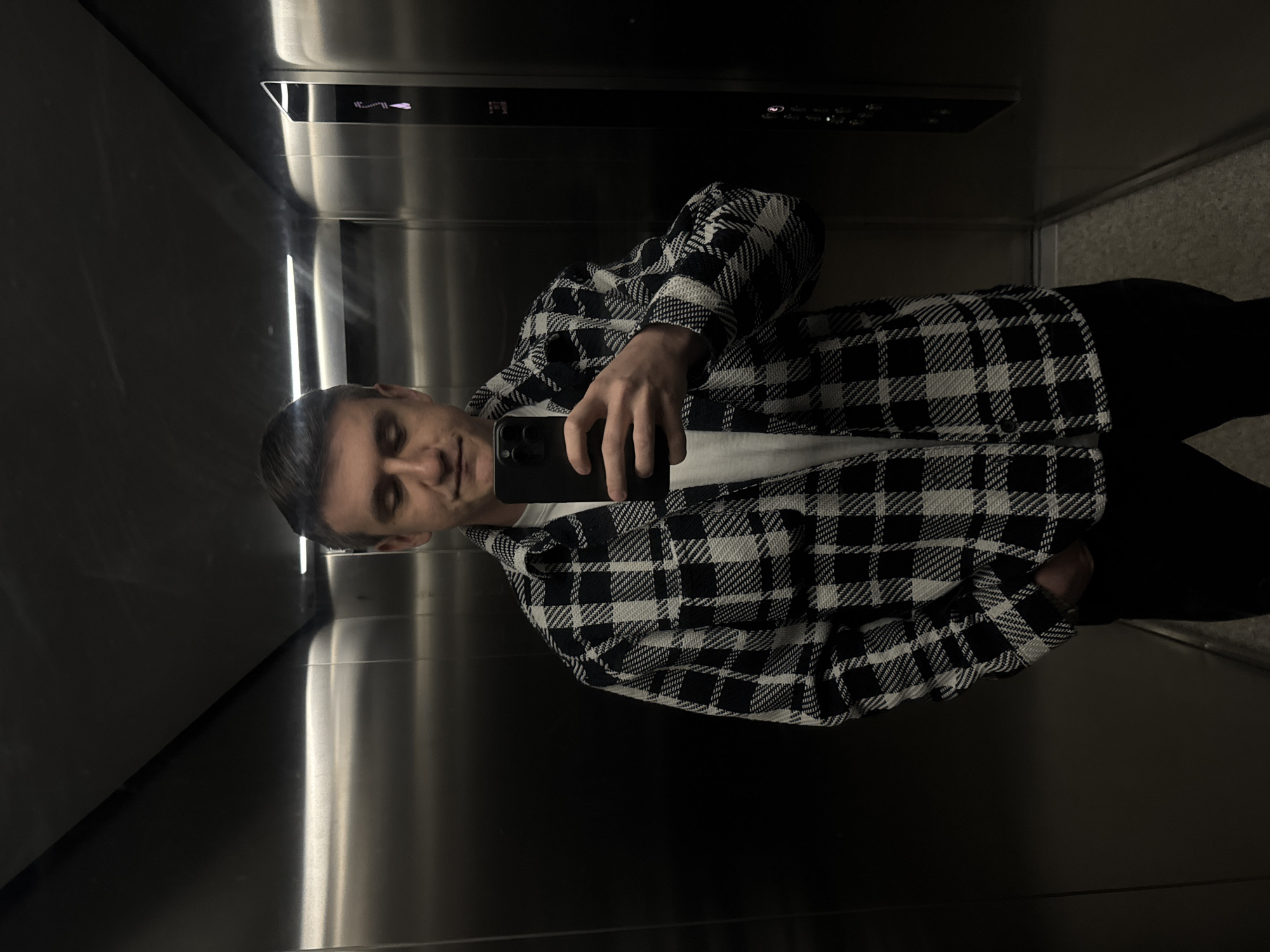 The image captures a selfie of a young man inside an elevator. He is positioned slightly to the left of the frame, seen from the chest up, holding a black smartphone to take the picture. He has short, dark hair and is wearing a black and white checkered jacket or thick shirt over a white undershirt, paired with dark trousers. His expression is neutral, with his eyes appearing somewhat closed or looking downwards, common for someone concentrating on a smartphone screen.
The setting is an elevator interior with highly reflective, brushed metallic walls that create a slightly distorted reflection of the man and his surroundings. A vertical light source is visible reflecting on the left wall, adding a gleam to the metallic surfaces.
In the reflection on the back wall, an elevator control panel is visible. It features an illuminated display showing what appears to be a number (possibly '1' or 'I') followed by several horizontal lines and an upward-pointing arrow, suggesting elevator status or direction. Below this display, there are several rows of buttons, some of which are illuminated. The overall lighting in the elevator is dim, which enhances the metallic sheen of the walls.
The activity taking place is the man taking a selfie. The location context provided is Warsaw, Poland, though no specific visual cues within the image confirm this. The scene portrays a casual, everyday moment, likely while in transit.