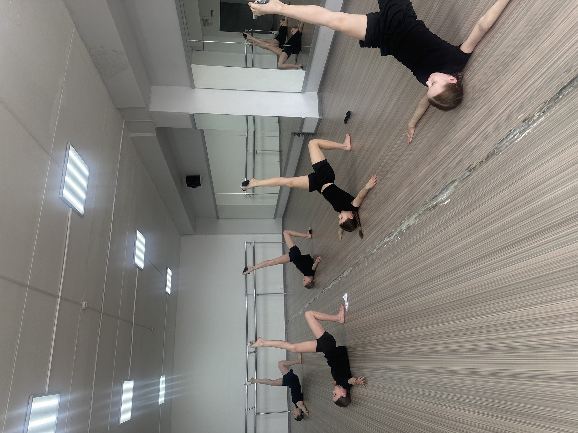 The image depicts a dance studio. Five young girls dressed in black outfits are practicing acrobatic poses, specifically a bridge with one leg extended vertically. They are arranged in a line stretching diagonally across the frame. In the background is a wall with a series of rectangular lights and a mirror reflecting the dancers. The floor is a striped, light-colored material. The location of the photo is a dance studio in Votkinsk, Russia.