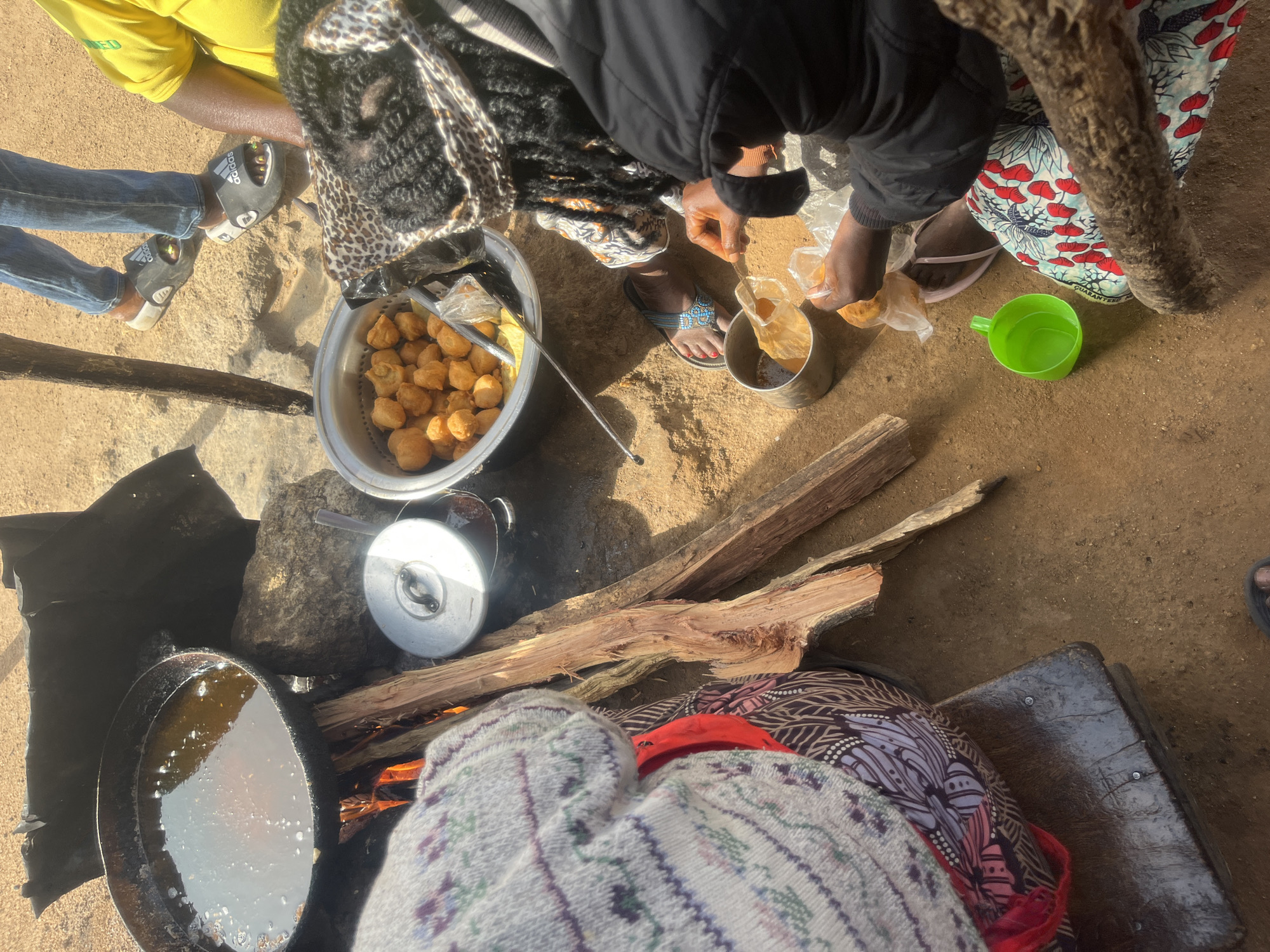 The image depicts an outdoor cooking scene in a sandy, outdoor environment, likely in Jos, Nigeria. The primary activity involves frying what appear to be fritters or dumplings in a large, dark pan over an open fire fueled by wood. The fire is visible beneath the pan, with flames licking upwards. To the right of the pan, a metal pot with a lid is placed on the ground.
Several individuals are present. In the foreground, a person wearing a patterned skirt and a black jacket is engaged in handling some of the cooked fritters, transferring them from a plastic bag into a small metal container. Another person, partially visible on the left, is wearing a patterned sweater and a red garment underneath, seated on a wooden stool. In the background, a person in blue jeans and Adidas slides stands near the cooking area. Another individual is partially visible on the far right, wearing a yellow shirt. A green plastic cup sits on the ground near the person handling the fritters. The overall scene suggests a communal food preparation activity in a simple, rustic setting.
