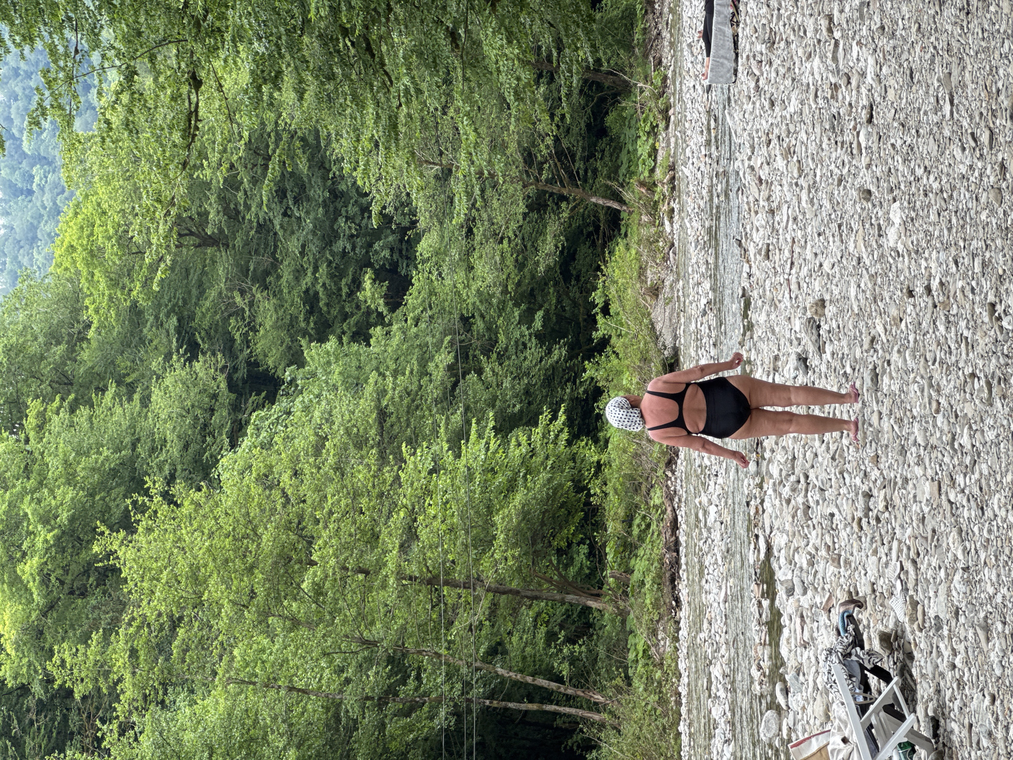 A person stands on a rocky riverbed, facing away from the camera, towards a shallow, clear river. The person is wearing a black one-piece swimsuit and a white patterned headscarf. The riverbed is covered in smooth, grey and white stones of various sizes. The river is shallow, with visible stones beneath the surface. On the left side of the image, there are some pieces of furniture and bags on the shore. The background is dominated by a dense, lush green forest that rises up a hill. Power lines are visible strung between two trees on the left side of the frame. The sky is overcast, creating a soft, diffused light. The scene appears to be a natural, undeveloped area, likely a riverbank in a forested region. The location is identified as Khleborob, Russia.