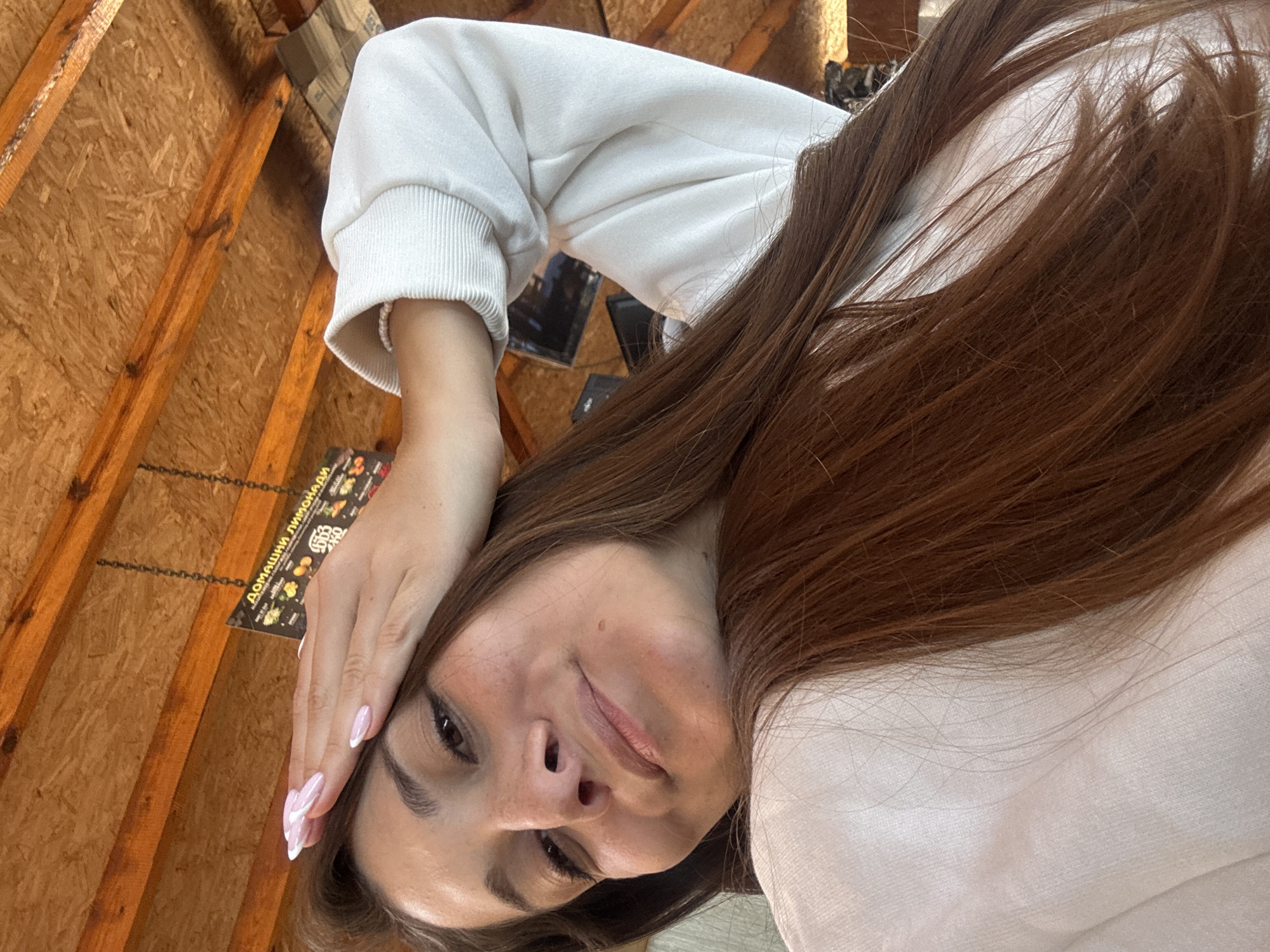 A selfie shows a woman with brown hair wearing a white sweatshirt, positioned at an angle against a backdrop of wood paneling and a framed menu. The menu features text in a language that may be Bulgarian, listing "Домашни лимонади" (Homemade Lemonades). The location is likely in Sozopol, Bulgaria, given the language and presumed location.