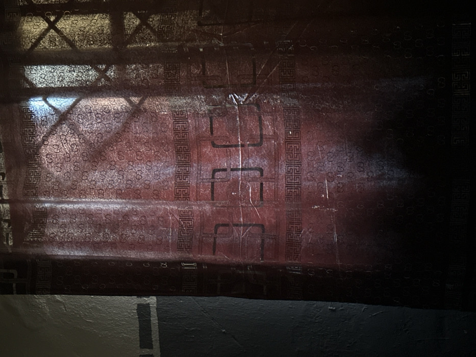 The scene depicts an interior view in Akwanga, Nigeria. A vertical, semi-translucent, red curtain is the central subject, featuring a repeating pattern of "GUCCI" logos interspersed with dark geometric border designs. The curtain material shows creases and minor surface wear. Light enters from an external source, illuminating portions of the curtain, with distinct angular shadows cast upon its surface, consistent with a window grille or security bars. To the left, a section of an interior wall is visible, painted in two horizontal bands: an upper lighter gray segment and a lower darker gray segment. No individuals, actions, or specific objects beyond the curtain and wall are discernible within the frame.
