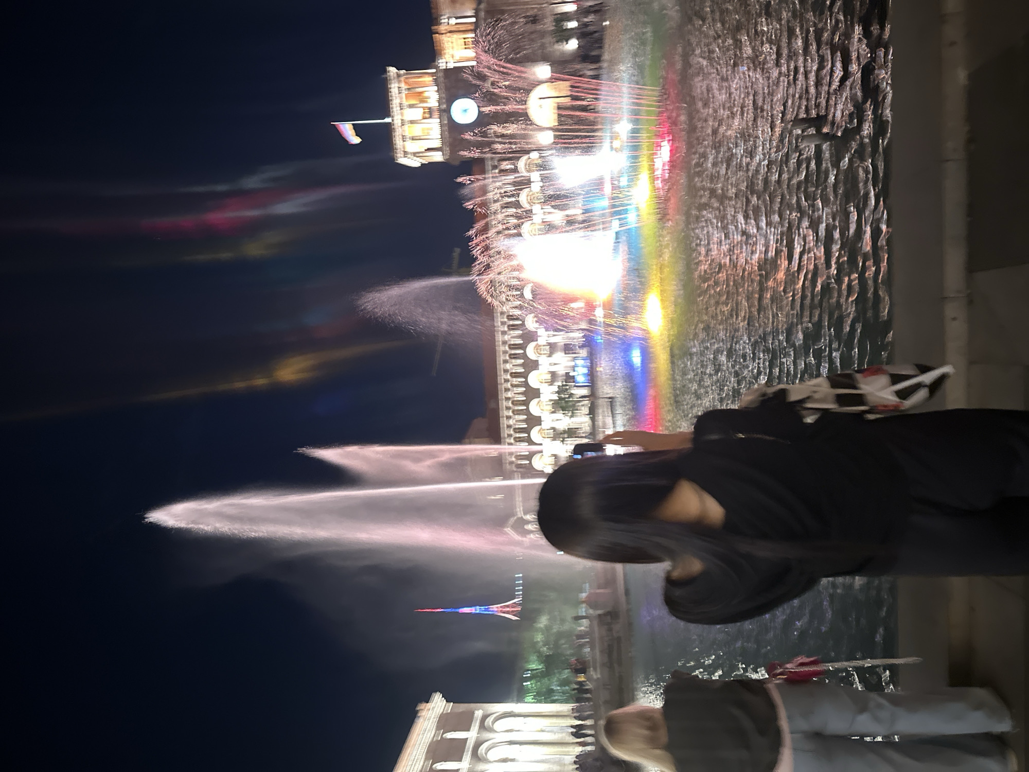 Captured at night, the image shows a fountain display in Yerevan, Armenia. A person with long, dark hair is partially visible in the foreground, seemingly observing the spectacle. Behind them, the fountain is illuminated with a rainbow of colors, with water jets reaching upwards. The water reflects the light, creating a vibrant, shimmering effect. In the background, a building with a clock tower and a flag on top stands tall. Additional individuals are visible in the lower portion of the frame, suggesting a public gathering.