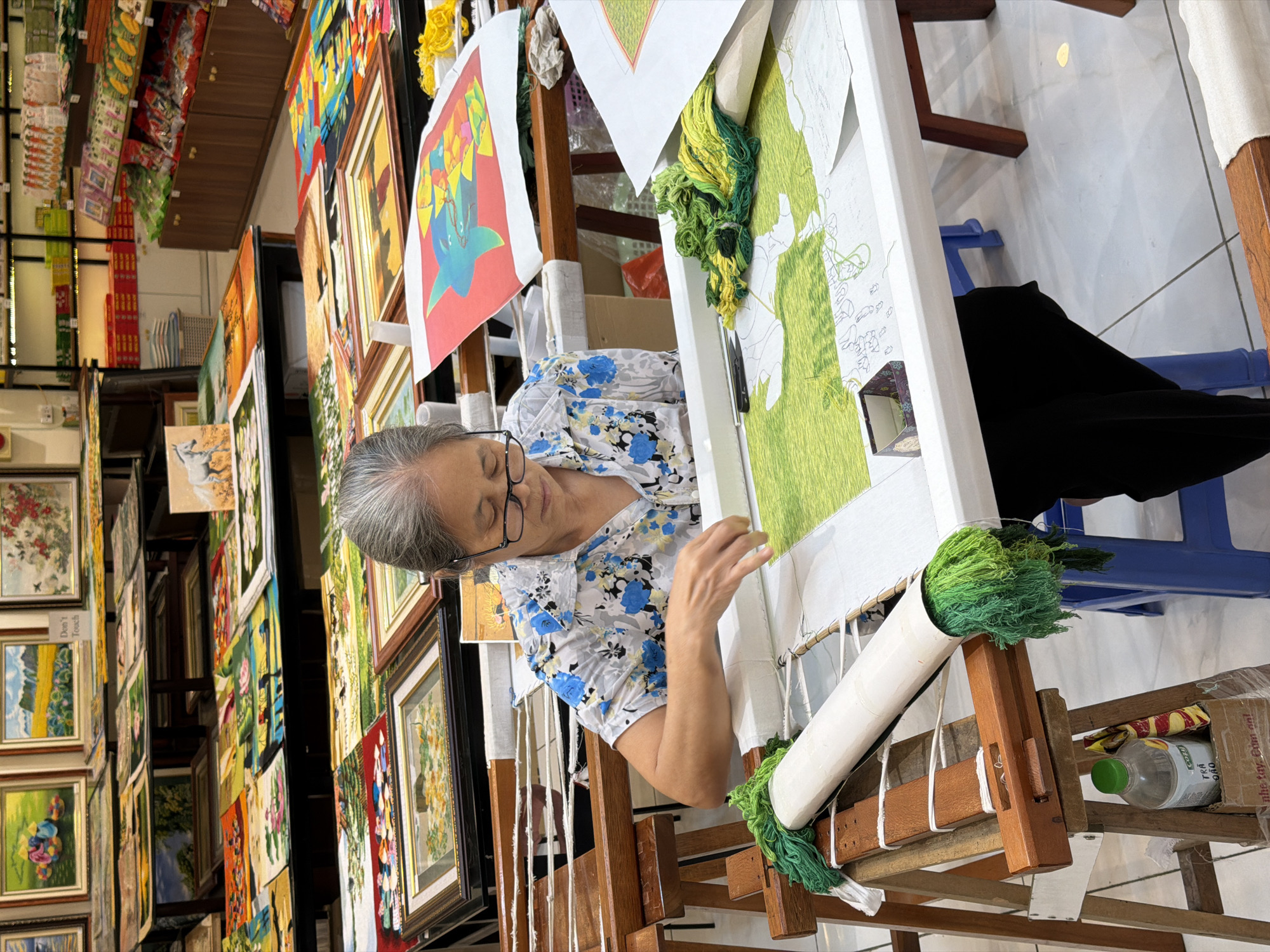 An elderly Vietnamese woman is depicted intently working on an intricate hand embroidery piece, likely in a studio or shop in Hải Phòng, Vietnam. She wears glasses and a white short-sleeved shirt with blue floral patterns, her expression one of focused concentration as she manipulates threads on a large wooden and white PVC pipe embroidery frame. The fabric on the frame shows a partially completed design in shades of green, with outlines for further work, and bundles of green and yellow threads hang from the frame. A colorful reference image is also attached.
The background is densely packed with numerous framed artworks, predominantly embroidered pieces, displayed on shelves and walls, creating a gallery-like setting. These artworks showcase various themes, including landscapes, flowers, and abstract patterns. A "Don't Touch" sign is visible on one of the displayed frames. Beneath the embroidery frame, a blue plastic stool provides seating, and a plastic bottle with the partial text "TRÀ ORO" and a box with "thể dục dụng cụ và" are visible. The scene captures the dedicated craftsmanship involved in traditional Vietnamese embroidery within a vibrant indoor environment, likely during daytime.