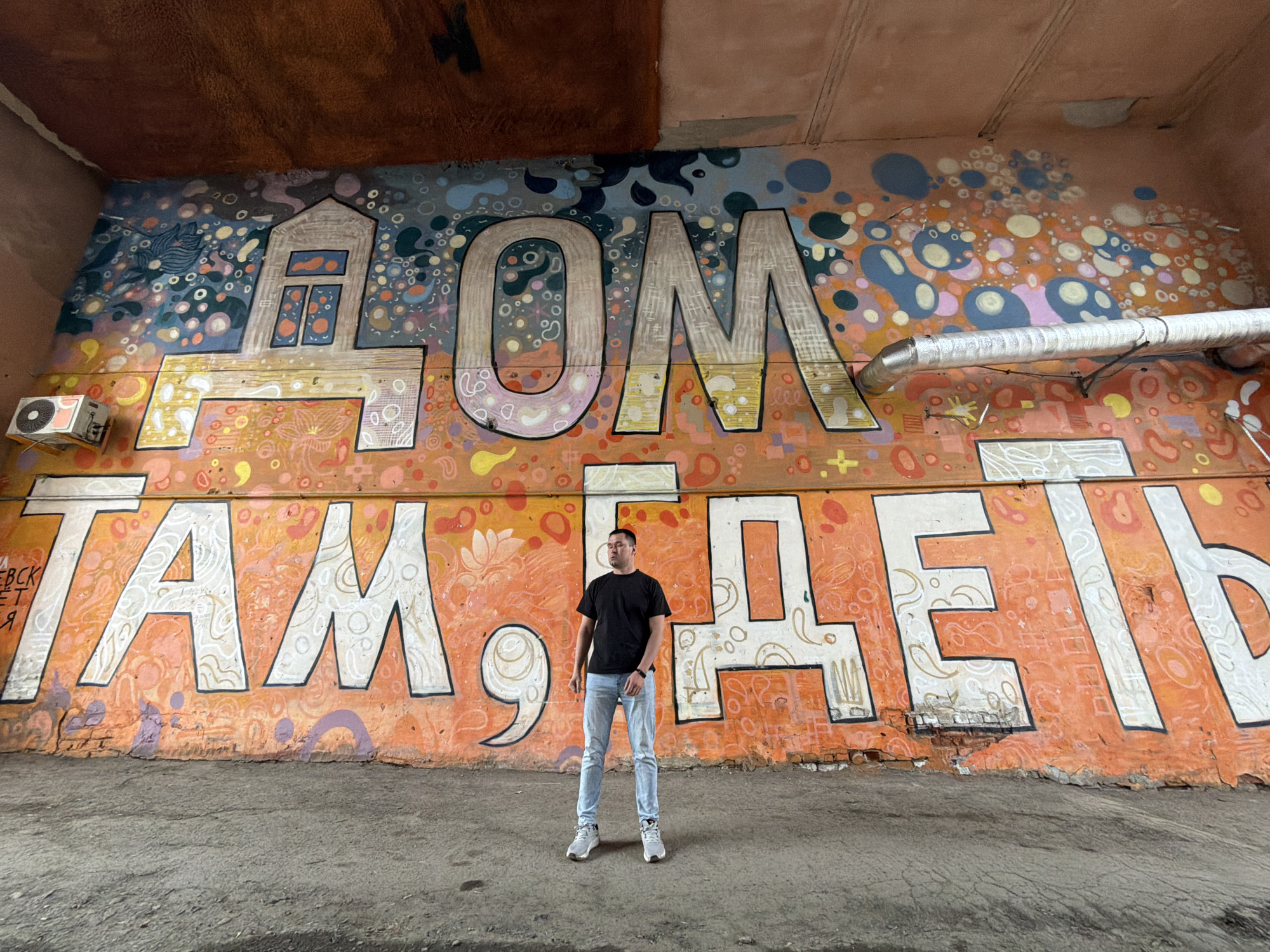 A full-body shot captures a man standing centrally in front of a large, colorful mural on a wall, beneath a concrete overpass. The setting is an urban outdoor environment, consistent with the provided location context of Izhevsk, Russia.
The man is dressed in a black short-sleeved t-shirt, light blue jeans, and light-colored sneakers. He has short, dark hair and stands with a neutral expression, looking slightly to his left (viewer's right).
The dominant feature of the scene is the vibrant mural, which covers most of the wall behind the man. It features a diverse palette of blues, greys, oranges, yellows, and pinks, adorned with abstract patterns of circles, bubbles, and organic shapes. Two prominent lines of large, white Cyrillic text with dark outlines are painted across the mural. The top line reads "ДОМ" (DOM), which translates to "HOUSE" or "HOME," with the initial letter 'Д' creatively stylized as a house with windows. The bottom line spells out "ТАМ ГДЕ Я" (TAM GDE YA), meaning "WHERE I AM," combining to form the phrase "Home is where I am." Smaller, less distinct Cyrillic text, possibly "НЕВСК" and parts of other words, is visible on the far left.
Above the mural, a rough, brownish-orange concrete ceiling, likely the underside of an overpass, spans the top of the image. An outdoor air conditioning unit is mounted on the wall to the left of the mural, and a silver-colored pipe runs horizontally across the upper right section. The ground below is cracked, grey concrete. The mural itself shows signs of wear, including some chipping and exposed brick along its lower edge. The bright, even lighting suggests it is daytime, possibly under an overcast sky or within the shade of the overhead structure.