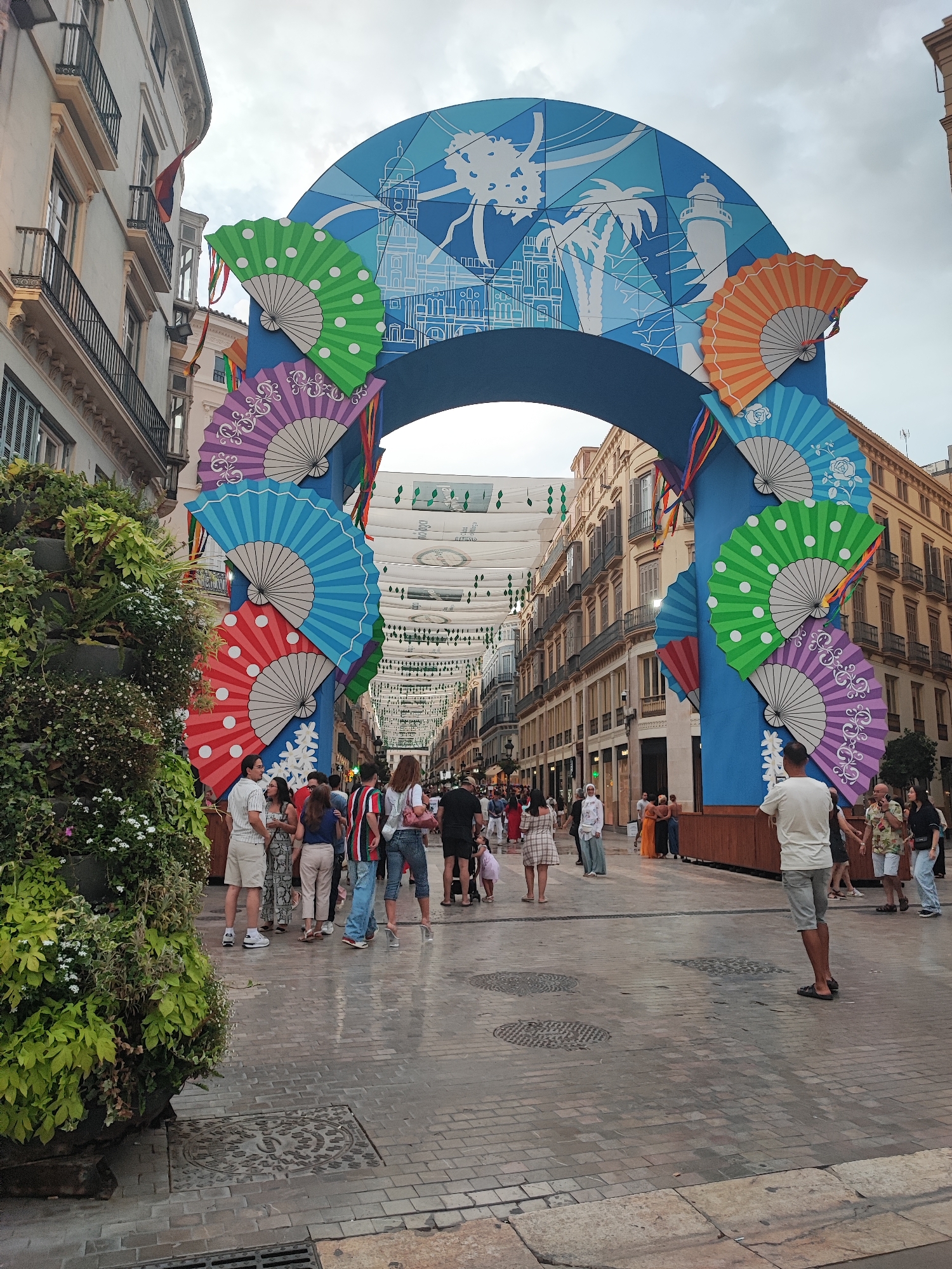 The image depicts a festive street scene in Málaga, Spain, likely during a celebration or festival. Dominating the center is a large, blue arched structure adorned with colorful paper fans in various shades of blue, green, purple, and orange. The arch itself is decorated with graphics featuring iconic Málaga landmarks such as the cathedral and a lighthouse, along with palm trees and geometric patterns. Suspended above the street are decorative banners with musical notes and what appear to be stylized lettering.
A crowd of people are gathered on the cobblestone street, some posing for photos, others walking or conversing. Among them are individuals dressed in casual summer attire. A prominent feature on the left side of the frame is a large, multi-tiered planter overflowing with lush green foliage and small white flowers, adding a natural element to the urban setting. The buildings flanking the street are multi-story structures with traditional balconies and architectural details. The sky is overcast, suggesting it is either early morning, late afternoon, or a cloudy day. The overall atmosphere is one of lively public gathering and celebration.