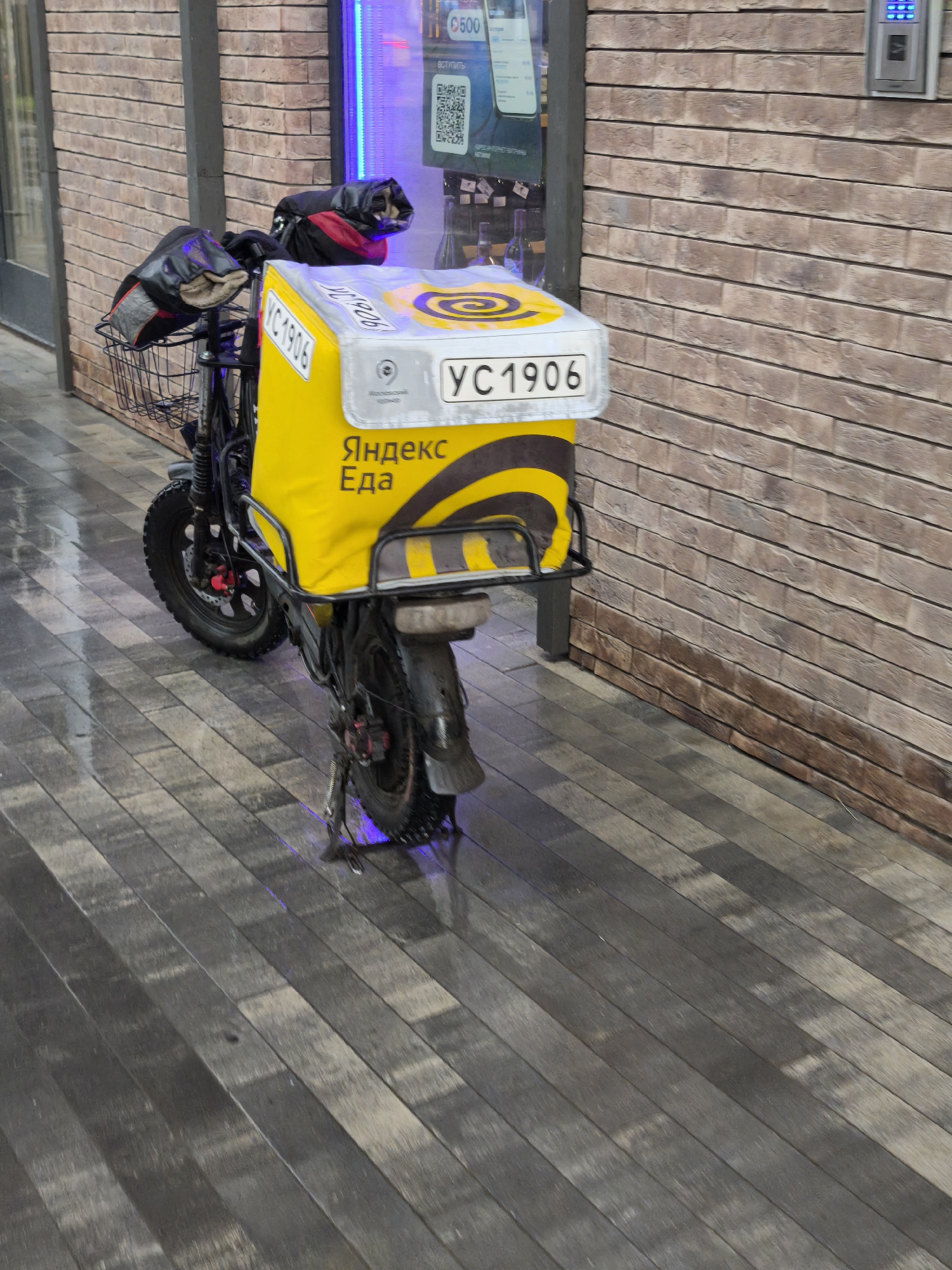 The image shows a food delivery bicycle in Moscow, Russia. The bicycle is parked on a wooden-planked walkway next to a brick building. It has a yellow "Yandex Eats" insulated delivery bag attached to the back with the license plate "YC1906". The bicycle has a front basket, and black gloves are placed on the handlebars. The environment appears to be wet.