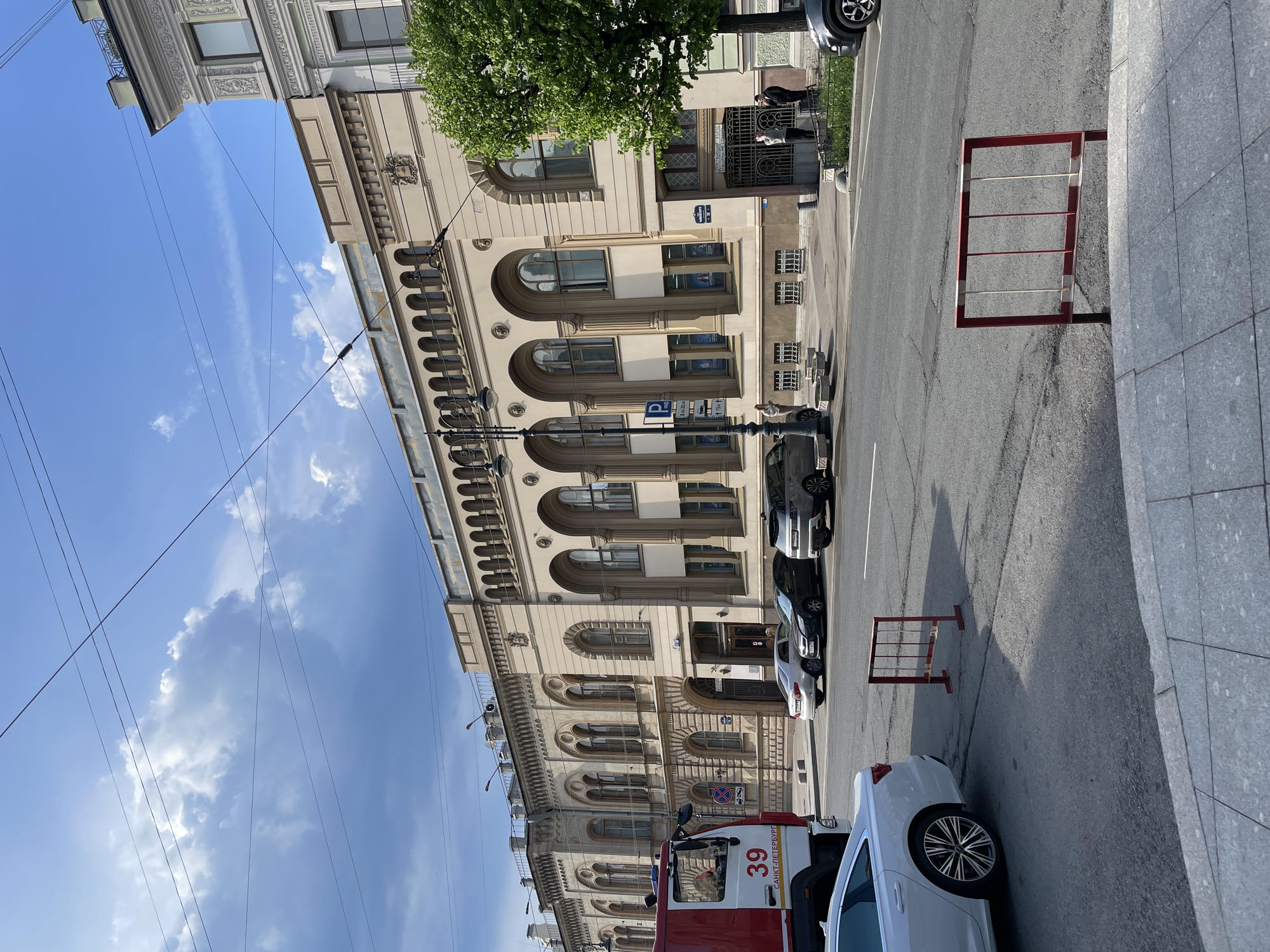 The image depicts a street in Saint Petersburg, Russia, on a partly sunny day. The architecture of the building on the left is notable, featuring arched windows and ornate detailing. A fire engine with "39 Санкт-Петербург" (Saint Petersburg) printed on its side is visible in the foreground. Several parked cars line the street. Two red and white safety barriers are placed on the street. Overhead, power lines stretch across the sky. People are standing at the entrance of the building.