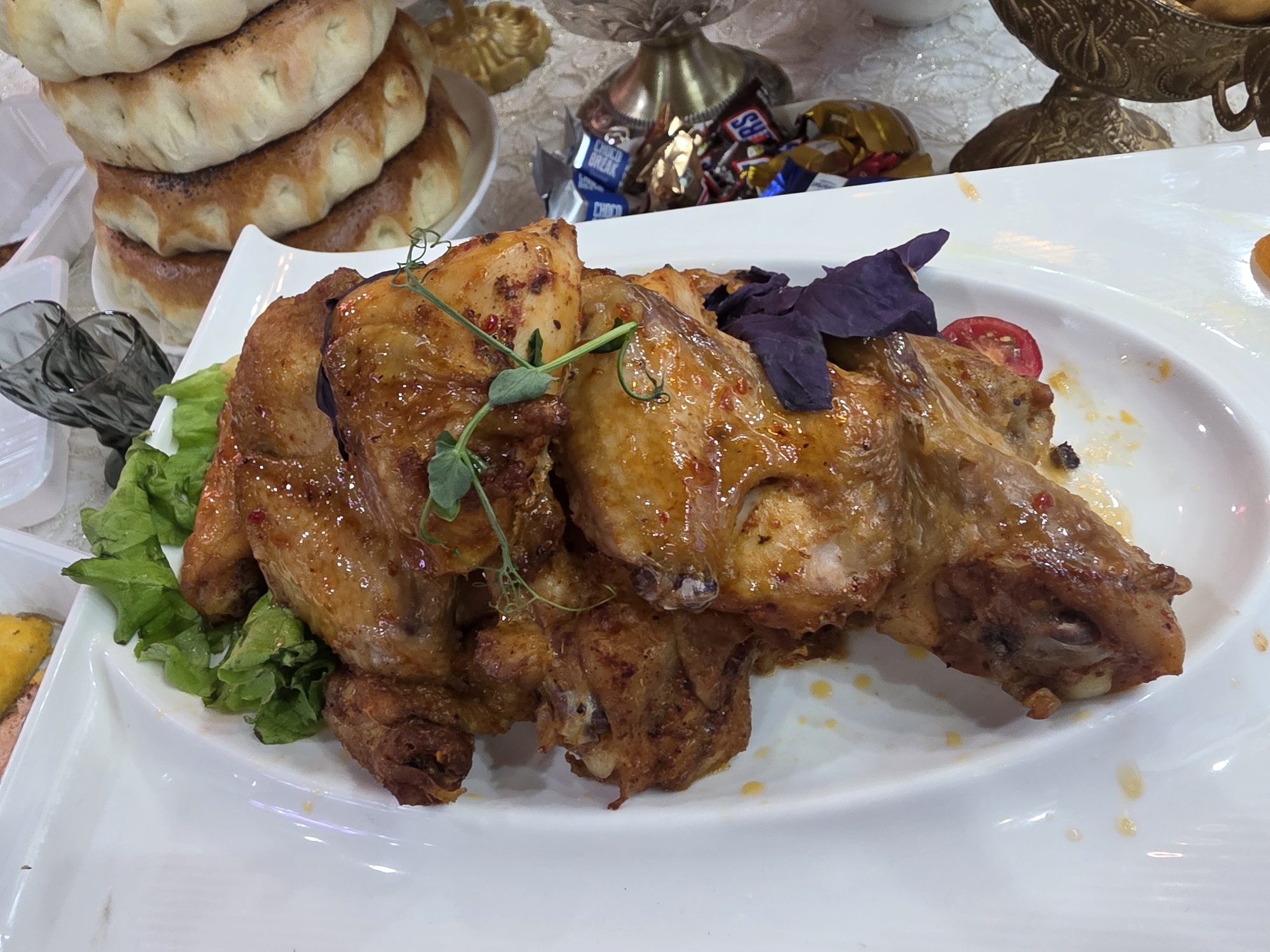 The image depicts a spread of food on a white table. In the foreground, a white rectangular platter presents a serving of roasted chicken, garnished with lettuce, purple basil, and sliced cherry tomatoes. To the left, a stack of round flatbreads sits on a plate. In the background, confectionery, including chocolate bars, rests near an ornate metal dish. The scene appears to be indoors and is located in Bishkek City, Kyrgyzstan, based on the visual cues of the food items and presentation styles commonly associated with the region.