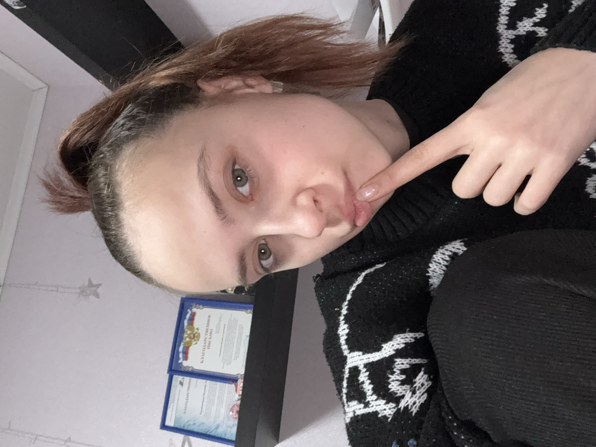 A young woman with fair skin and light-colored eyes is pictured indoors, likely in a home setting. Her reddish-brown hair is pulled back, and she is wearing a black sweater with a white, snowflake-like pattern. Her right index finger is touching her lower lip. Behind her, there is a shelf with two framed certificates. The certificates feature text in Russian and an emblem bearing the colors of the Russian flag, suggesting the scene is likely in Russia. Based on the text on the certificates, the location may be Прикамский. The wall is painted in a light color, adorned with a decorative star string light.