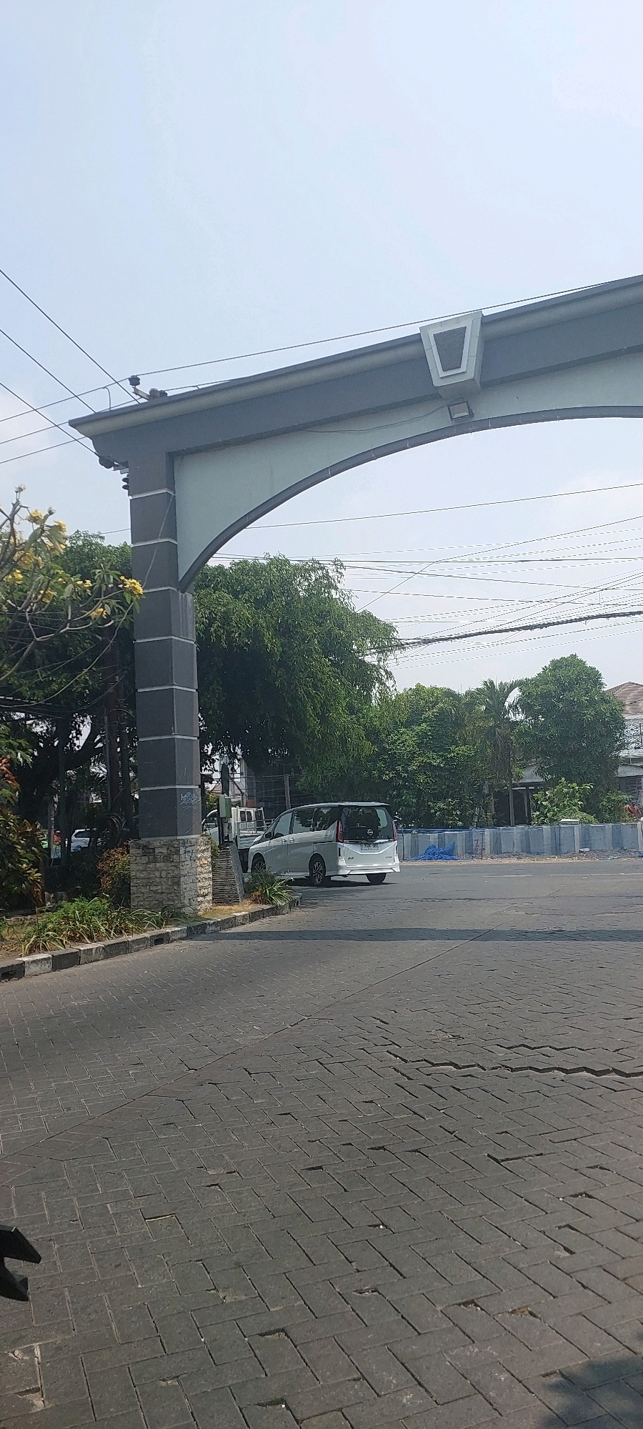 The image depicts an arched gateway with a dark grey rectangular support pillar on the left, topped with a light grey archway and horizontal beam. A silver minivan is parked in front of the pillar. The road surface is paved with dark grey rectangular bricks laid in a herringbone pattern. Green trees and buildings are visible in the background, with power lines crisscrossing the sky. The scene appears to be on a sunny day. The location is identified as a residential area entrance in Surabaya, Indonesia.