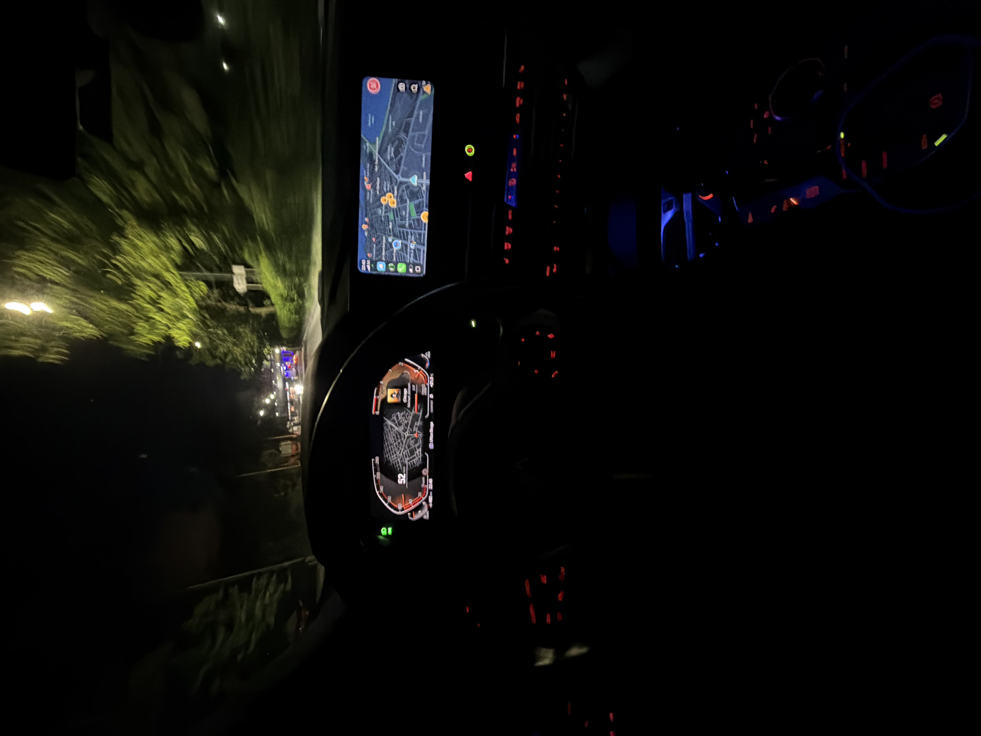 The image is a first-person view from inside a car at night, traveling on a street lined with trees. The car's dashboard is illuminated, showing a digital speedometer indicating 52. A GPS map is displayed on the central infotainment screen. The interior of the car has red ambient lighting. Outside the car, streetlights and the headlights of other vehicles are visible. The overall scene suggests a nighttime drive through a moderately lit urban or suburban area.